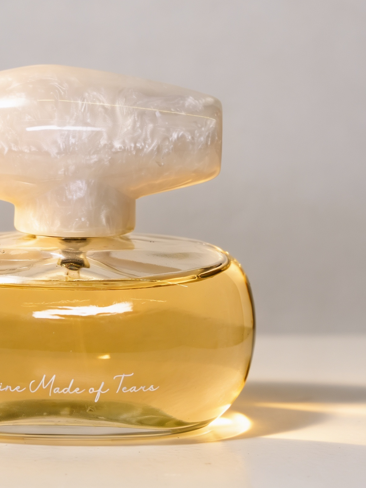 Close-up of a glass perfume bottle with a pearl-colored cap. The bottle contains light amber-colored perfume. The text on the bottle says 'Made of Tears'.