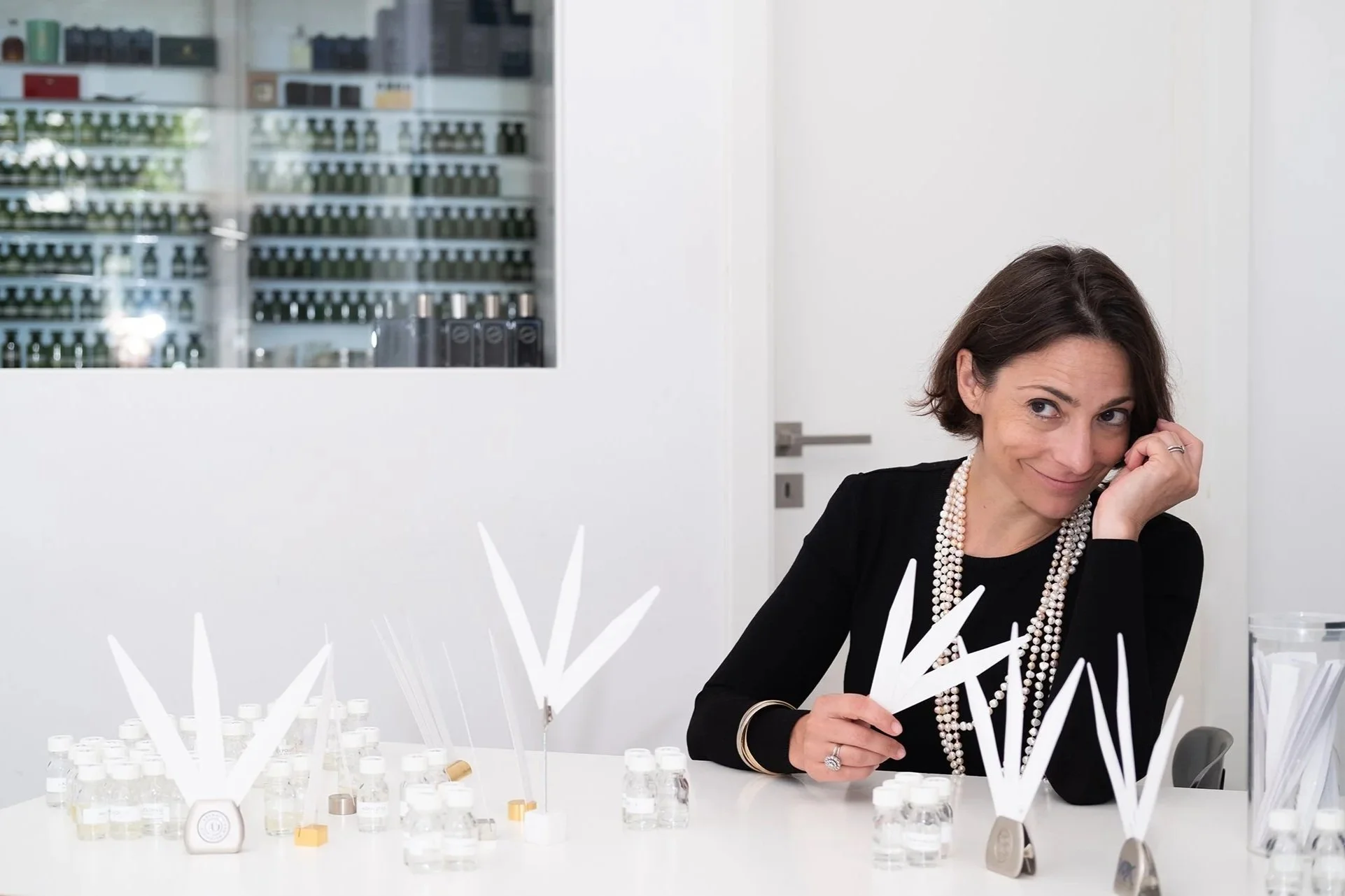 A smiling woman with short dark hair, wearing a black top and a pearl necklace, is sitting at a table with perfume test strips.
