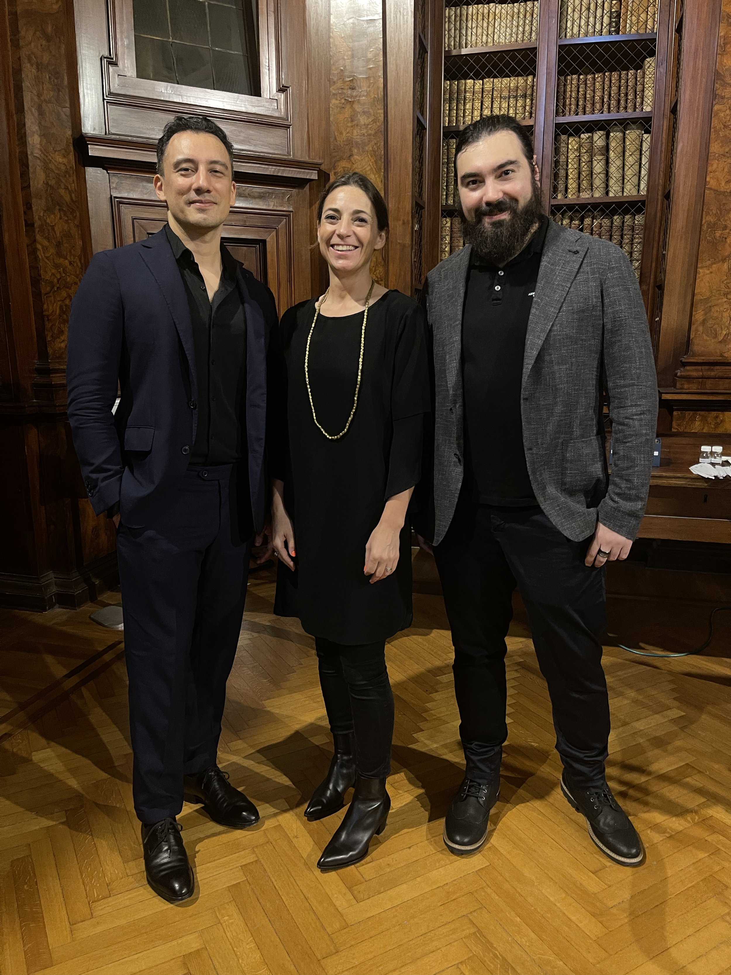Three people standing in a room with wooden walls and bookshelves. A man in a dark suit is on the left, a woman in the middle is smiling, and a man with a beard in a gray blazer is on the right. All are dressed in dark clothing.