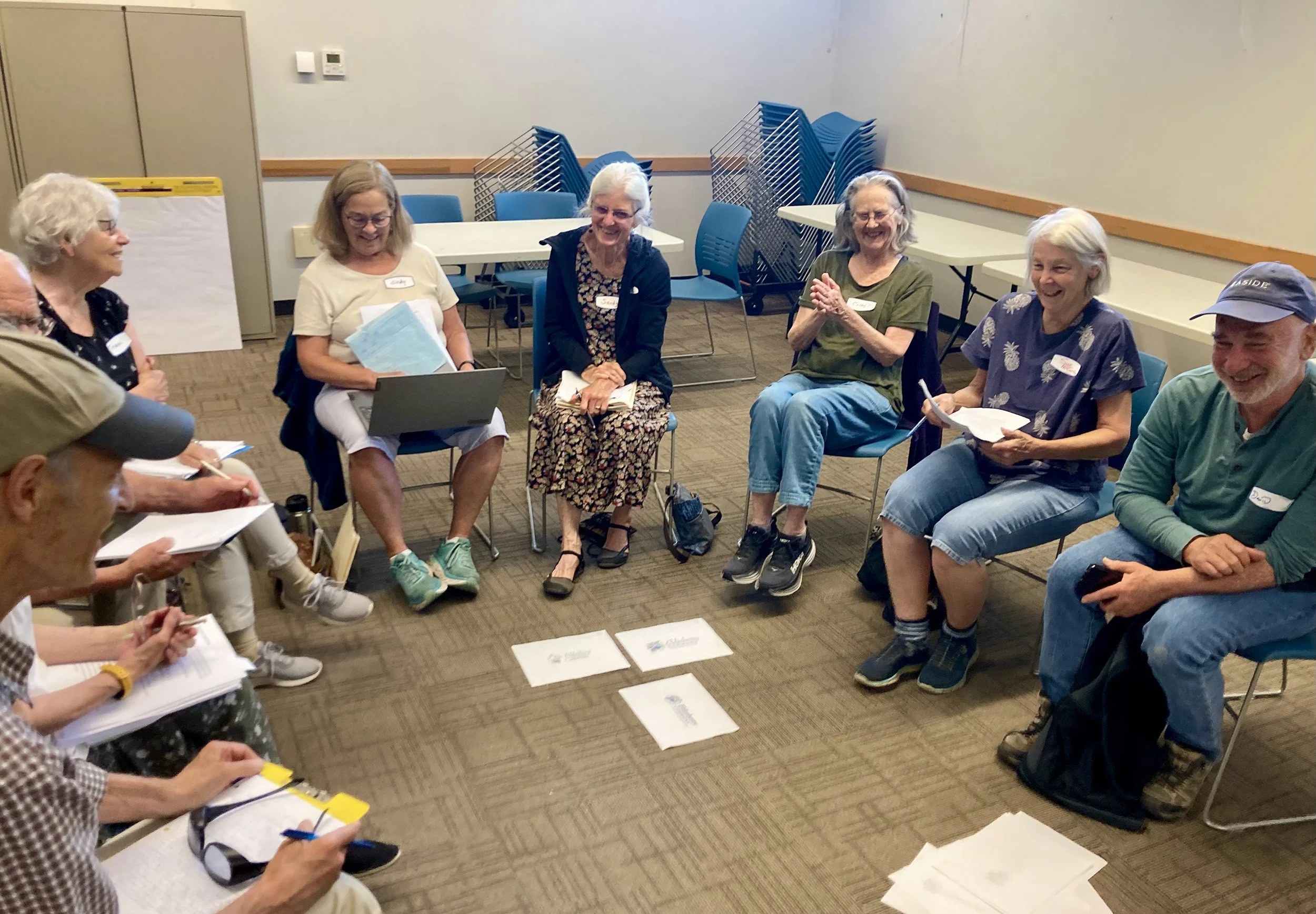 Group of elderly people sitting in a circle in a meeting room, smiling and engaging in discussion, with papers and a laptop visible.