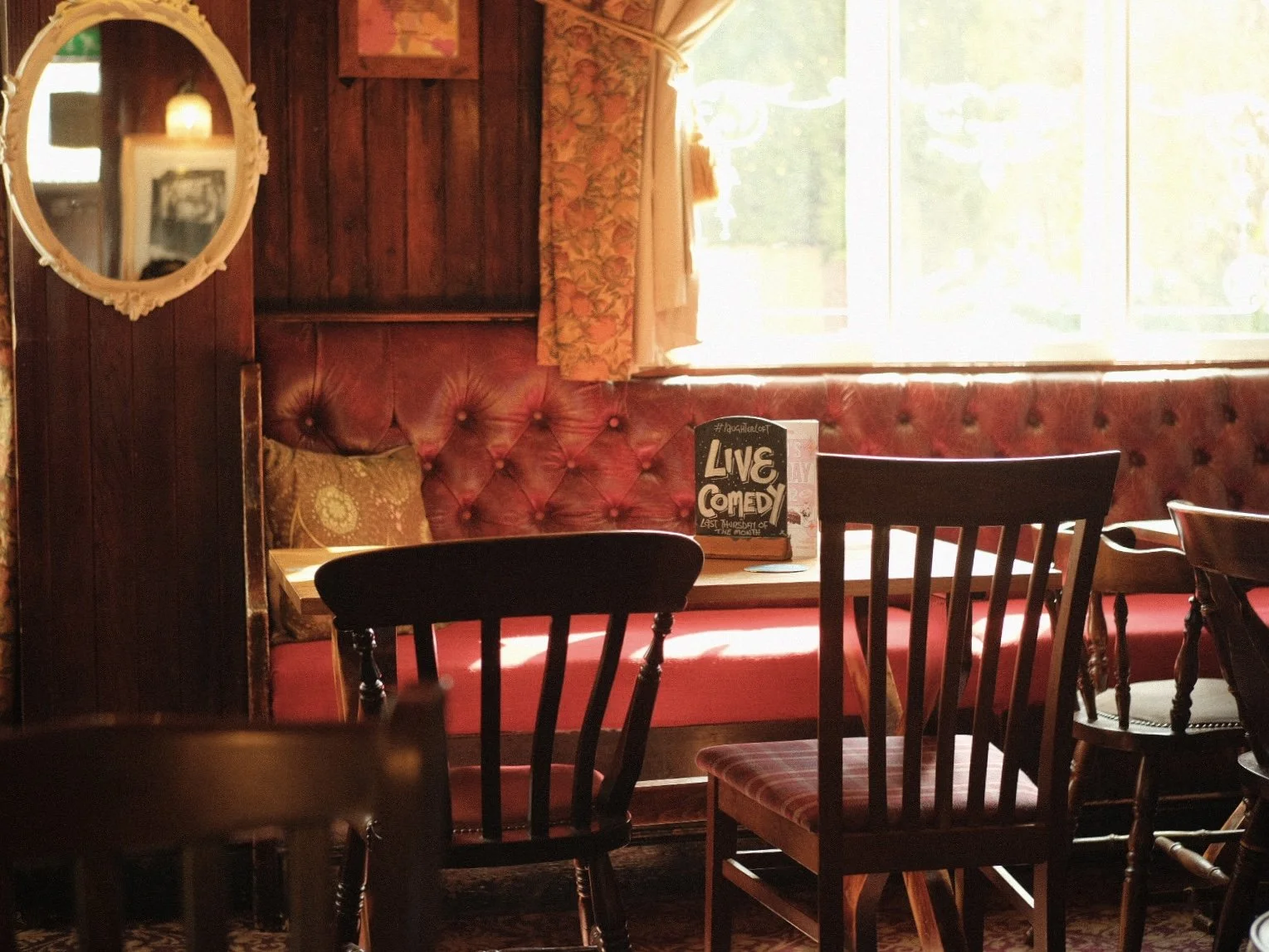 Interior of a cozy pub or restaurant with wooden walls, red tufted upholstery, and chairs around tables. A window lets in natural light, and a sign on a table reads 'Live Comedy.'
