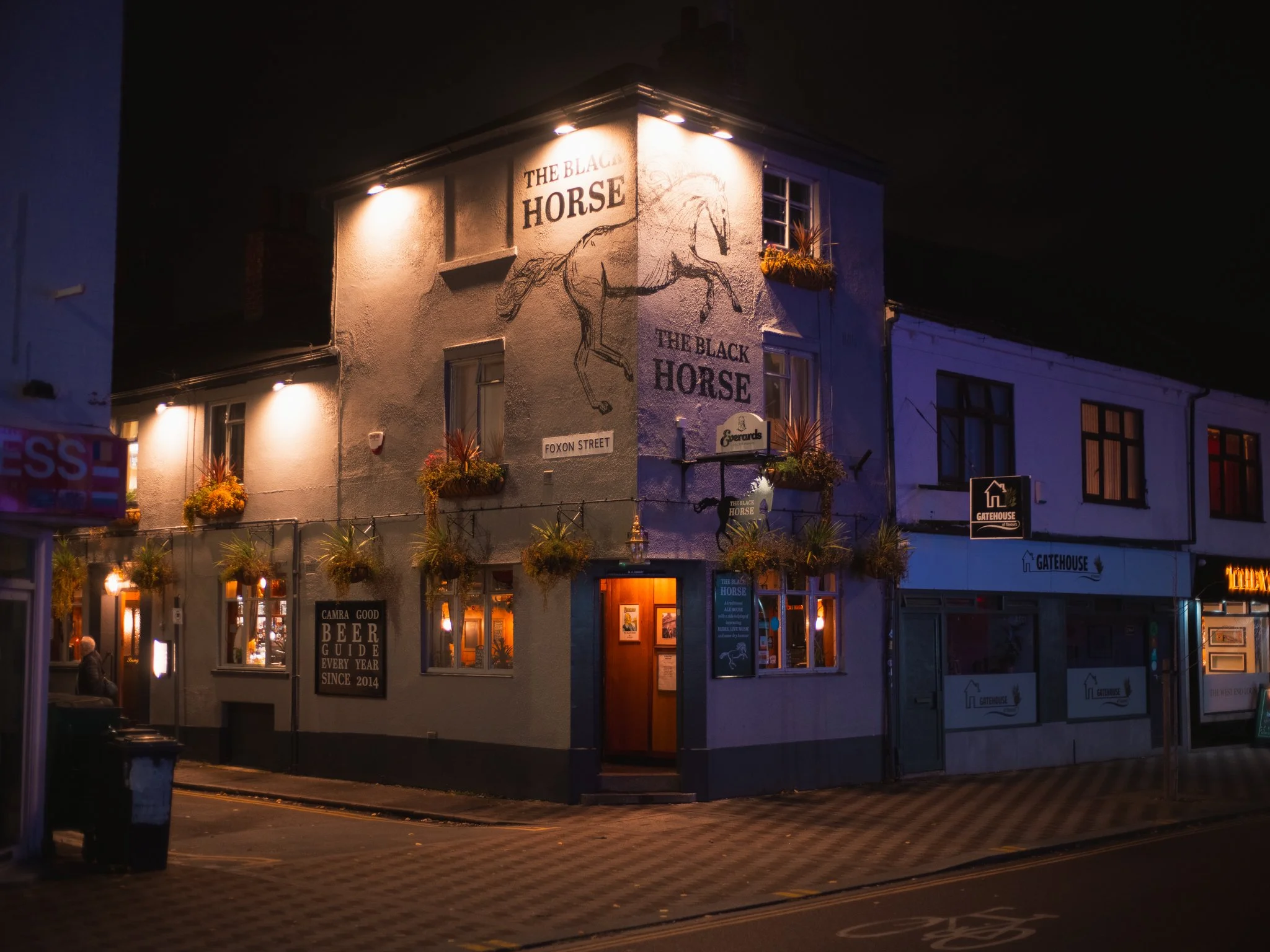 A night view of The Black Horse pub with painted horse mural, illuminated signs, hanging flower baskets, and lit windows on a street corner.