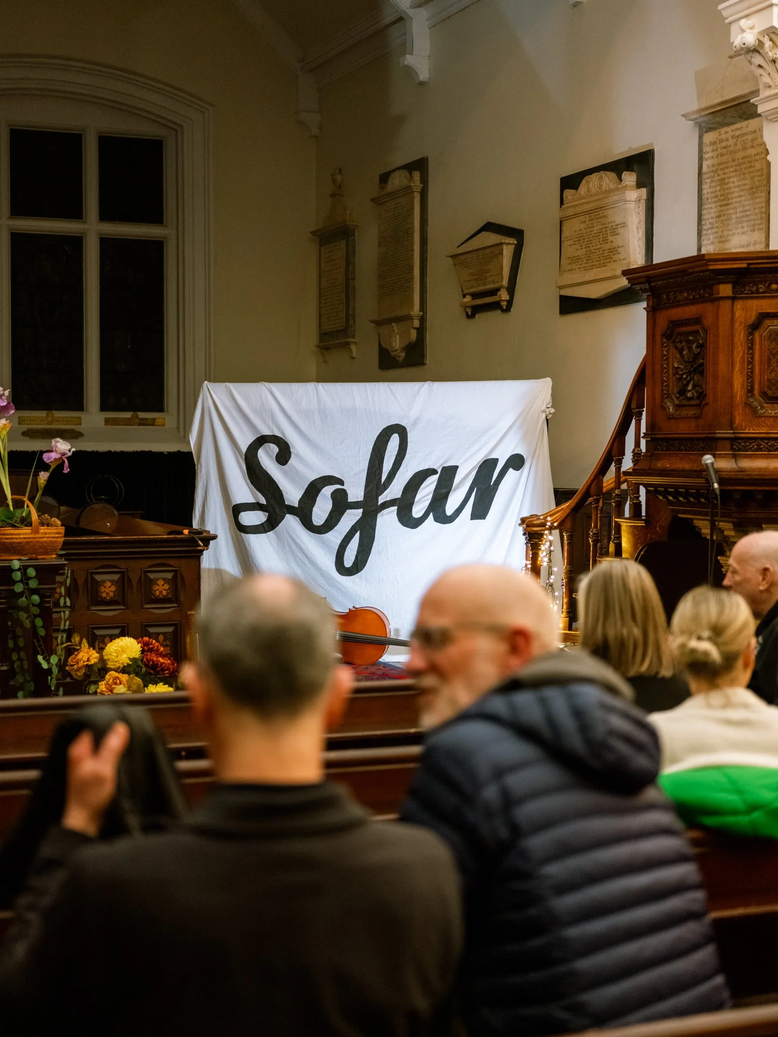 People seated in a church or concert hall watching a performance, with a large white cloth displaying the word 'Sofar' in black cursive lettering above the stage, which has a violin resting on a stand and flower arrangements.