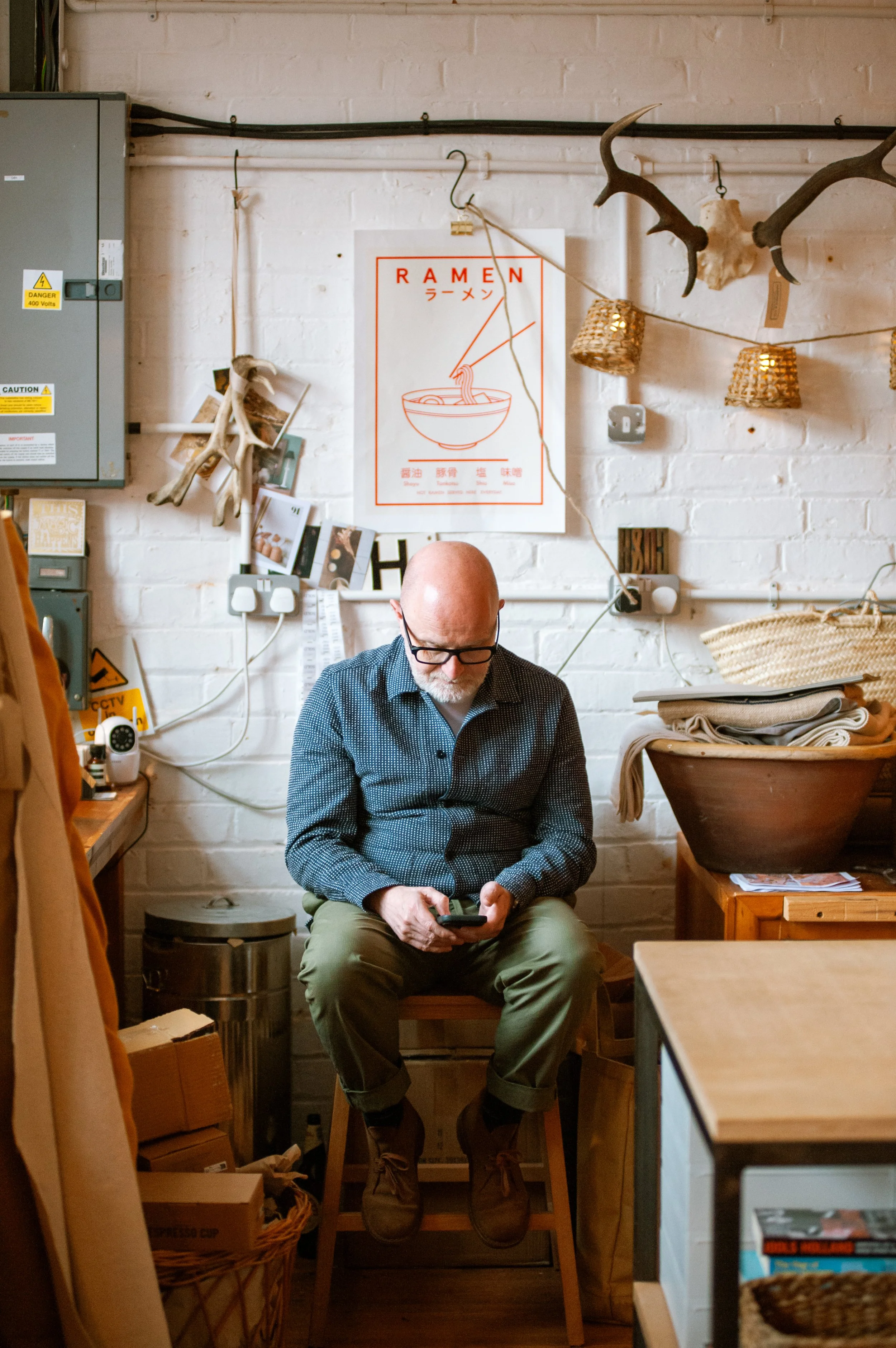 A man with glasses, a bald head, and a beard is sitting on a stool in a cozy, rustic room, looking at his phone. The room has a white brick wall decorated with a ramen poster, antlers, string lights, and various decor items. There are shelves with books and blankets, and the area appears warm and inviting.
