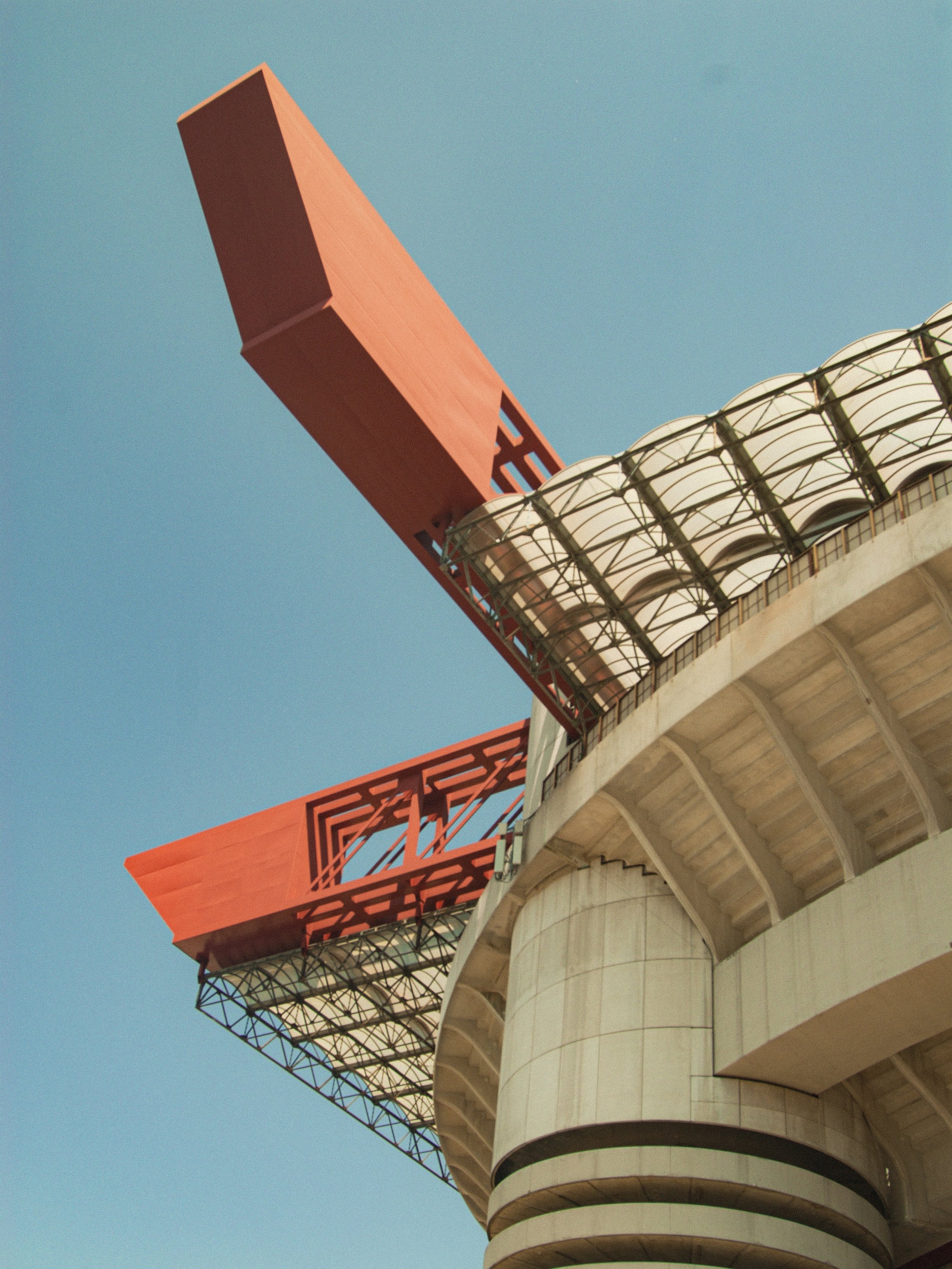 Close-up of a sports stadium's architectural details, showing orange structural elements extending from a curved, concrete building with seating and glass roof panels, against a clear blue sky.