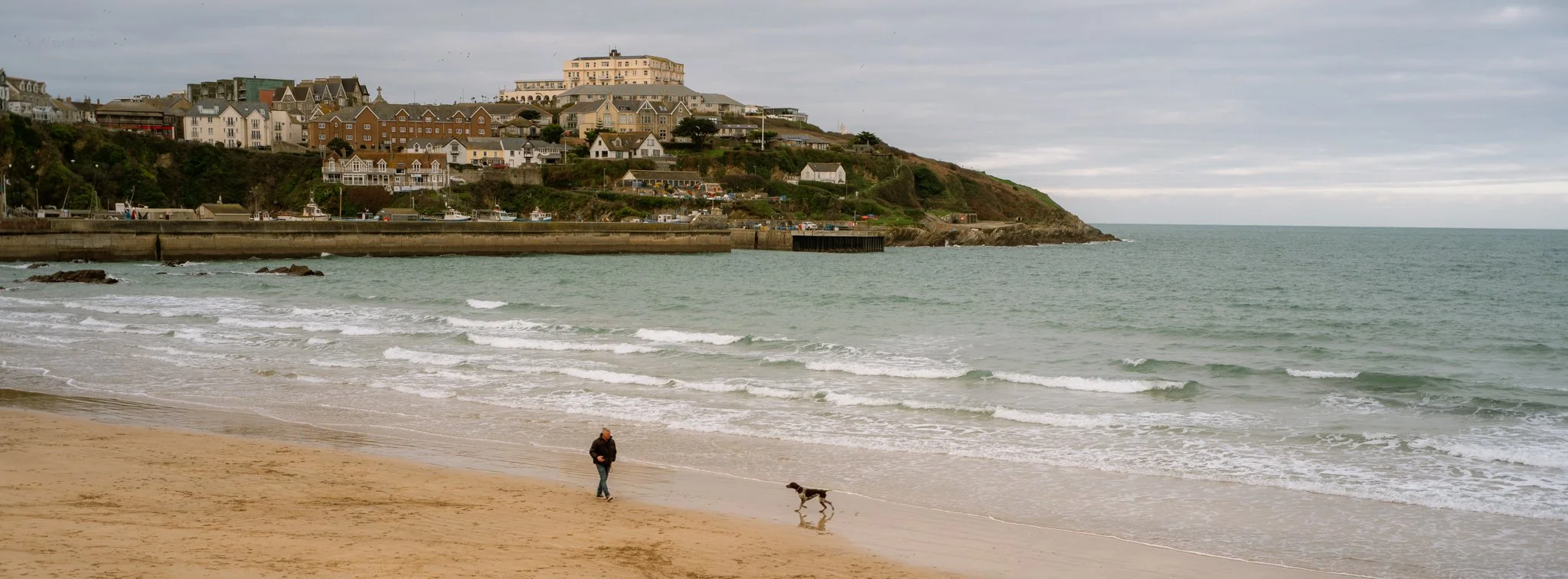 Person walking a dog along the sandy beach with waves and a coastal town on a hill in the background under a cloudy sky.