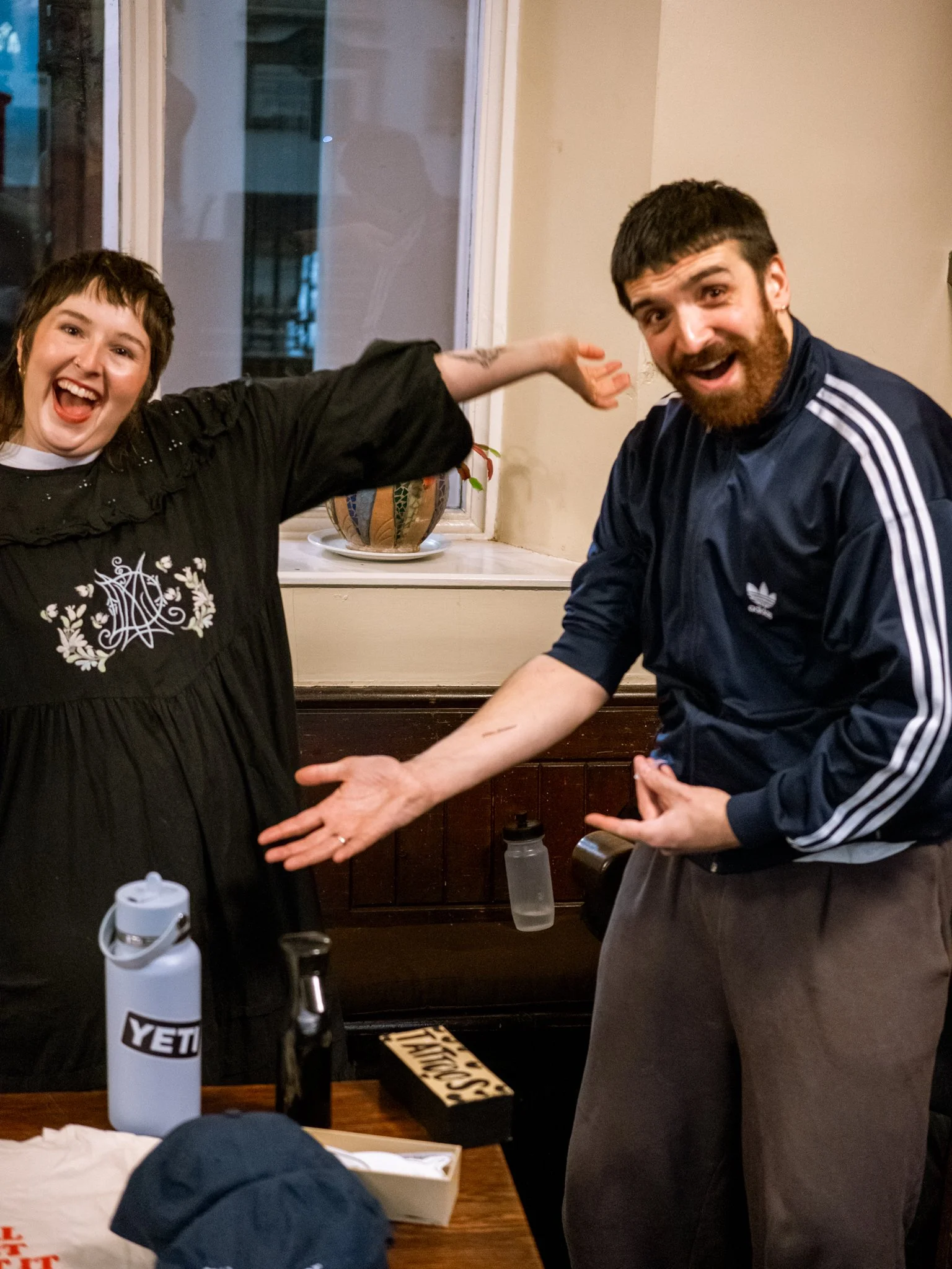 Two people are celebrating at a restaurant or cafe, smiling and showing excitement, with one woman on the left pointing at the man on the right.