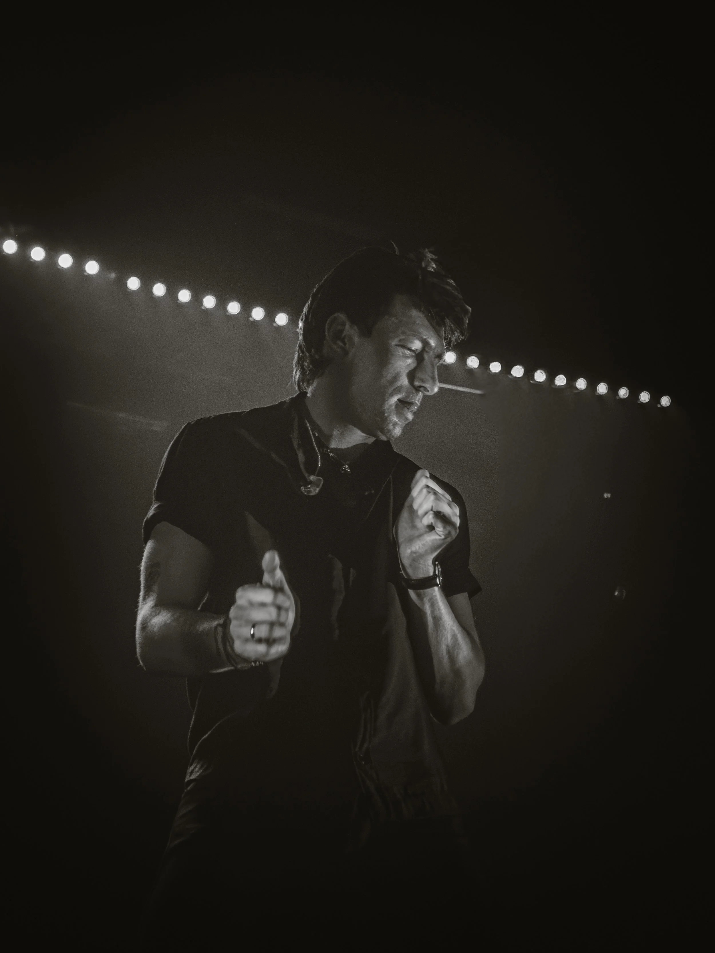 A man with dark hair and a beard singing into a microphone on stage, illuminated by stage lights in black and white.