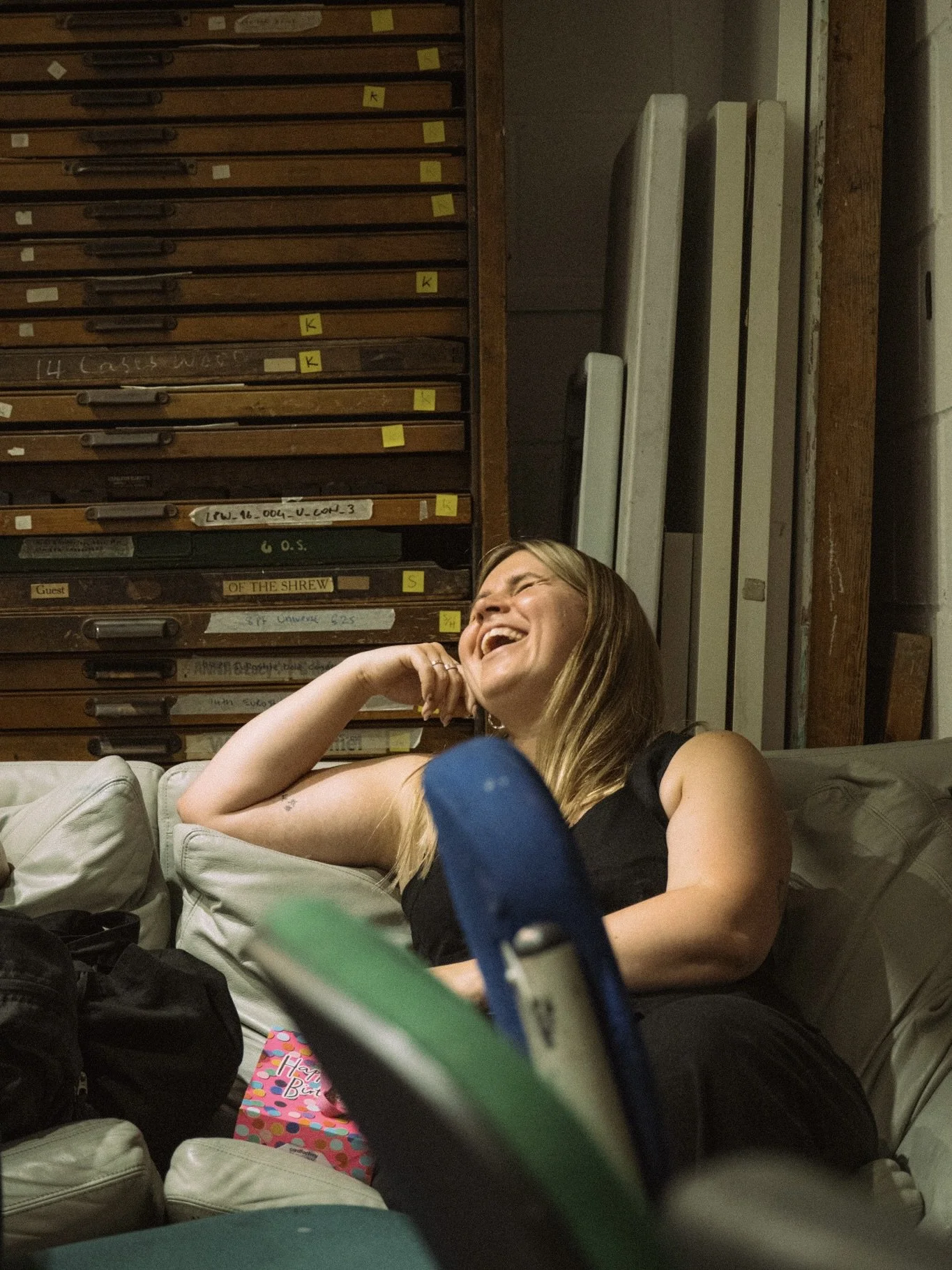 A woman with blonde hair laughing while reclining on a couch in an indoor setting with wooden drawers and metal objects in the background.