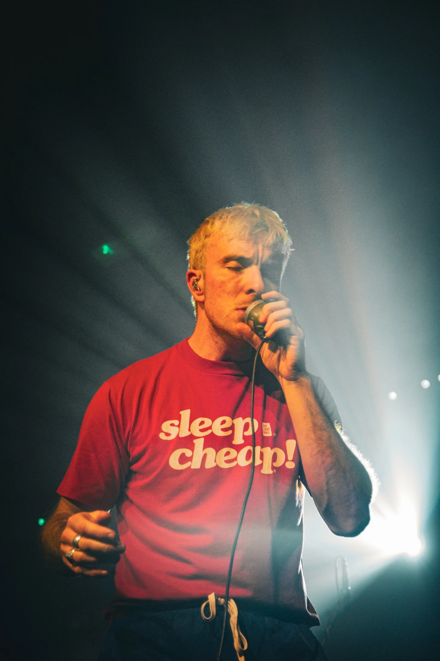 A young man with blonde hair singing into a microphone on stage, wearing a red t-shirt that says 'sleep cheap!'