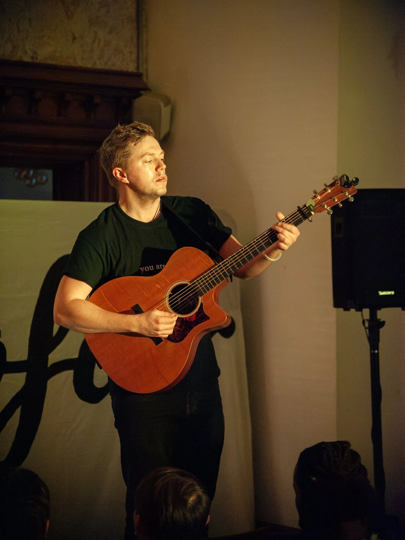 A young man playing an acoustic guitar on stage in front of an audience, with a speaker nearby and a partial view of the audience in the foreground.