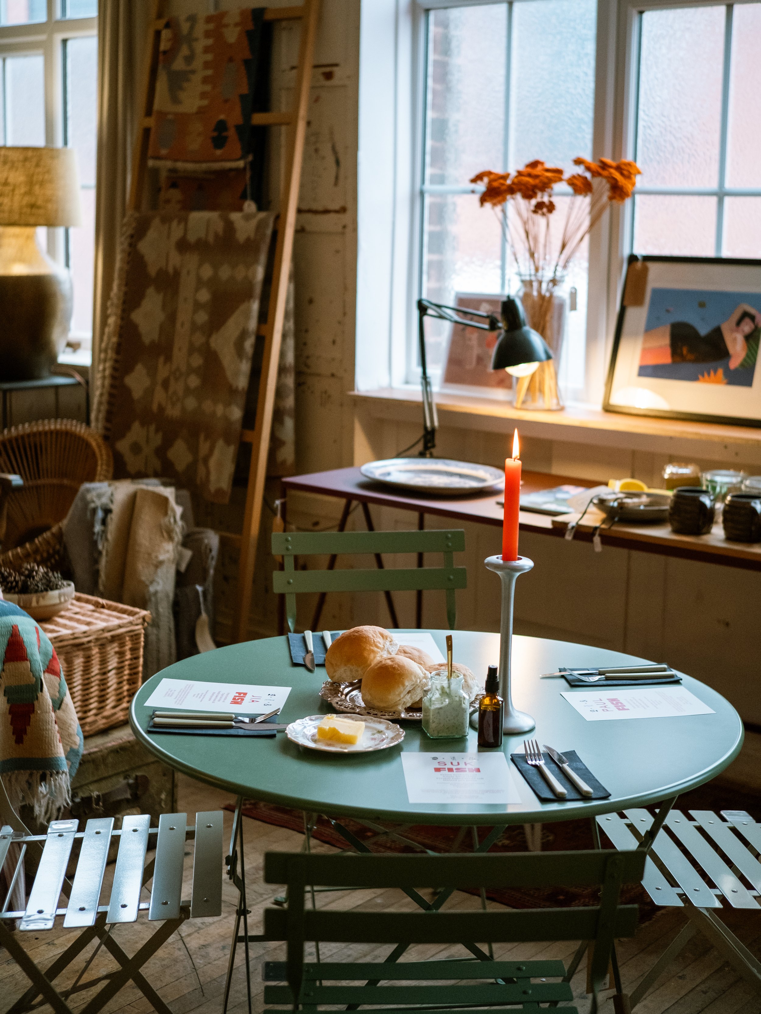 A cozy dining setup in a rustic room with a round green table, a red candle on a white candlestick, bread rolls, butter, and condiments, set for a meal. The background features a window with sunlight, dried flowers, artwork, and various decorative ob
