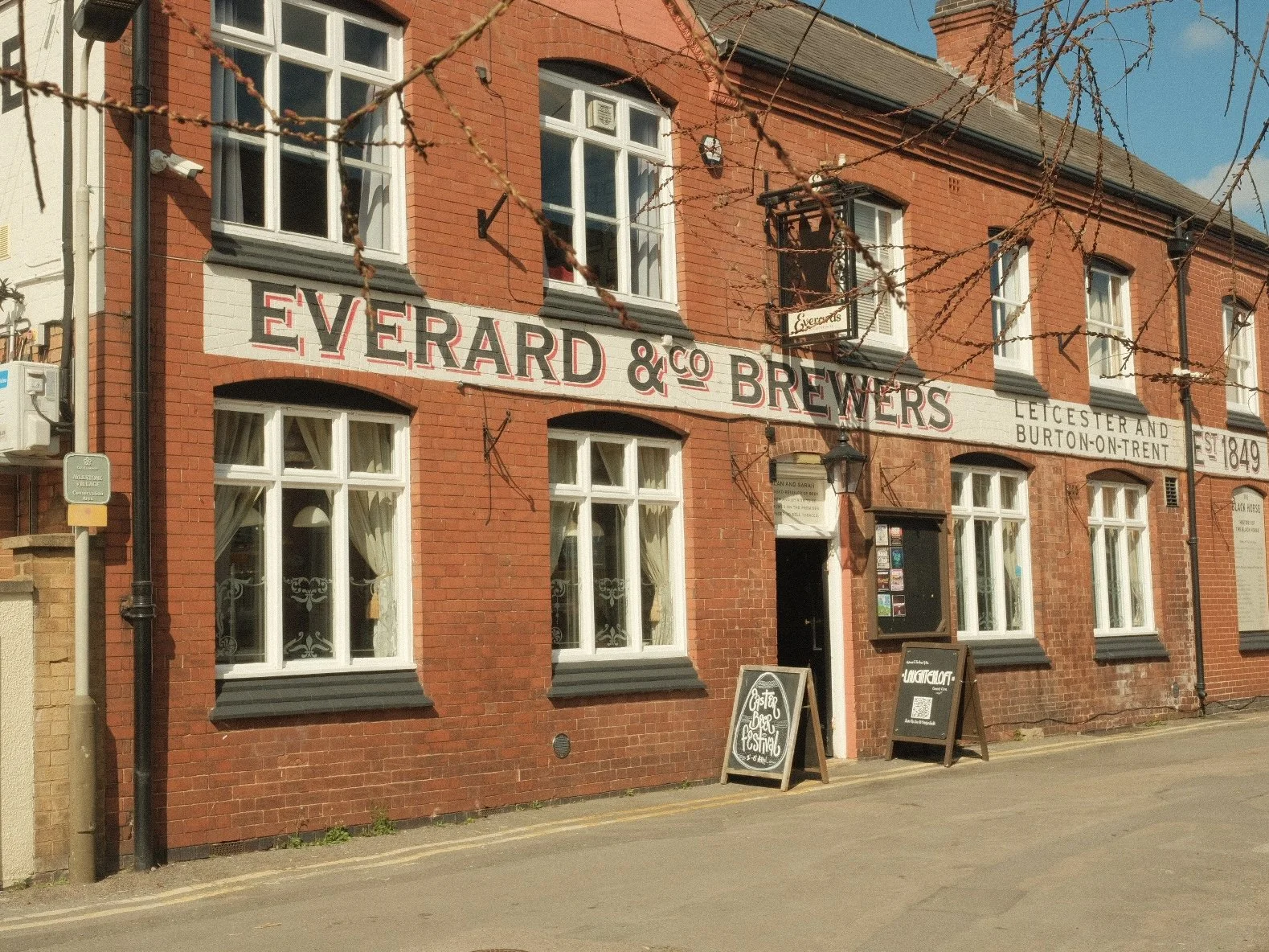 Red brick building with sign reading Everard & Co Brewers in Burton-on-Trent, established 1849. Windows with white curtains and outdoor chalkboard signs advertising Easter Beer Festival.