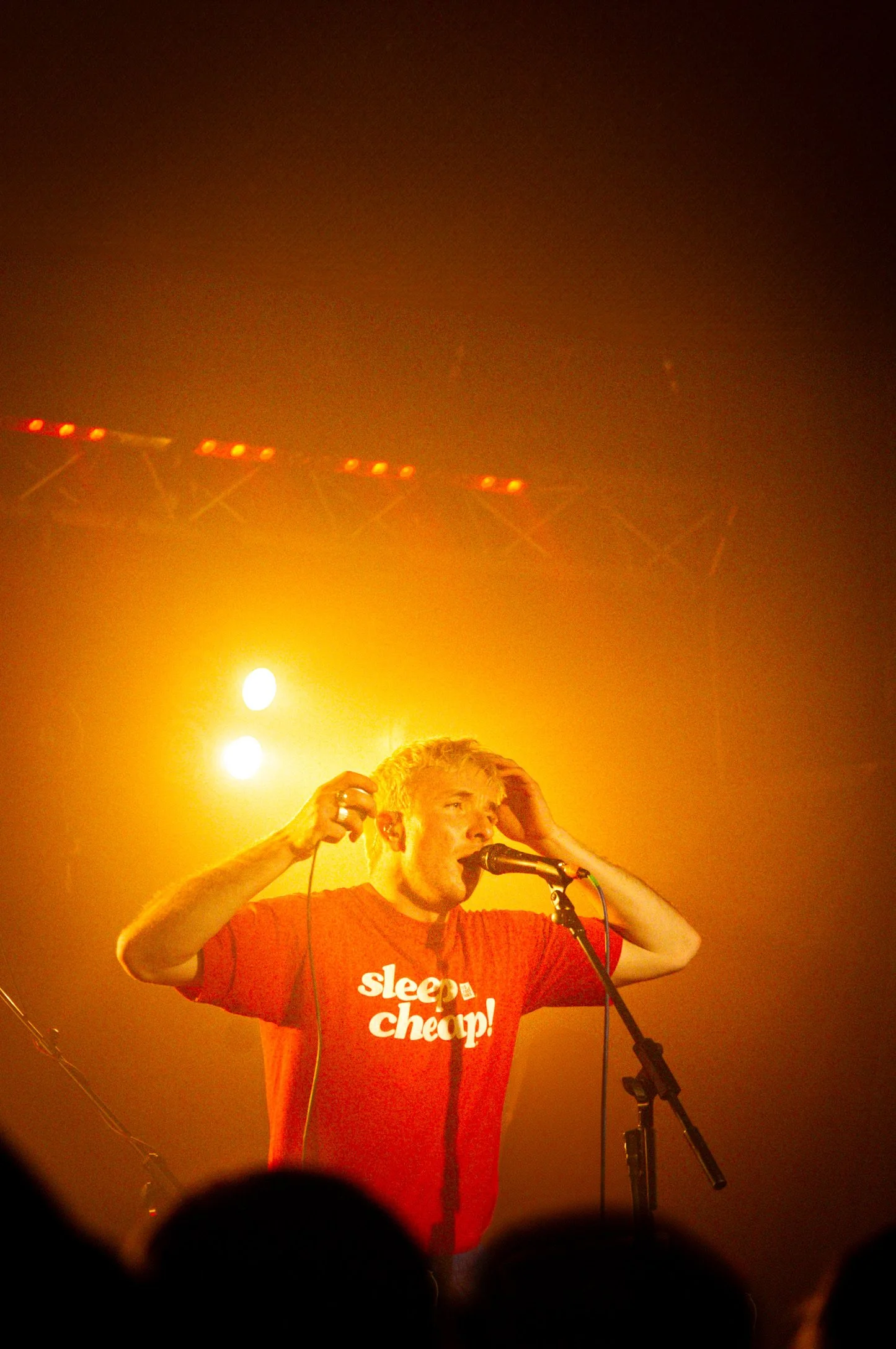 A male singer with platinum blonde hair, wearing a red t-shirt with the words 'sleep cheap!' on it, performing on stage under yellow lighting while holding a microphone and adjusting his hair.