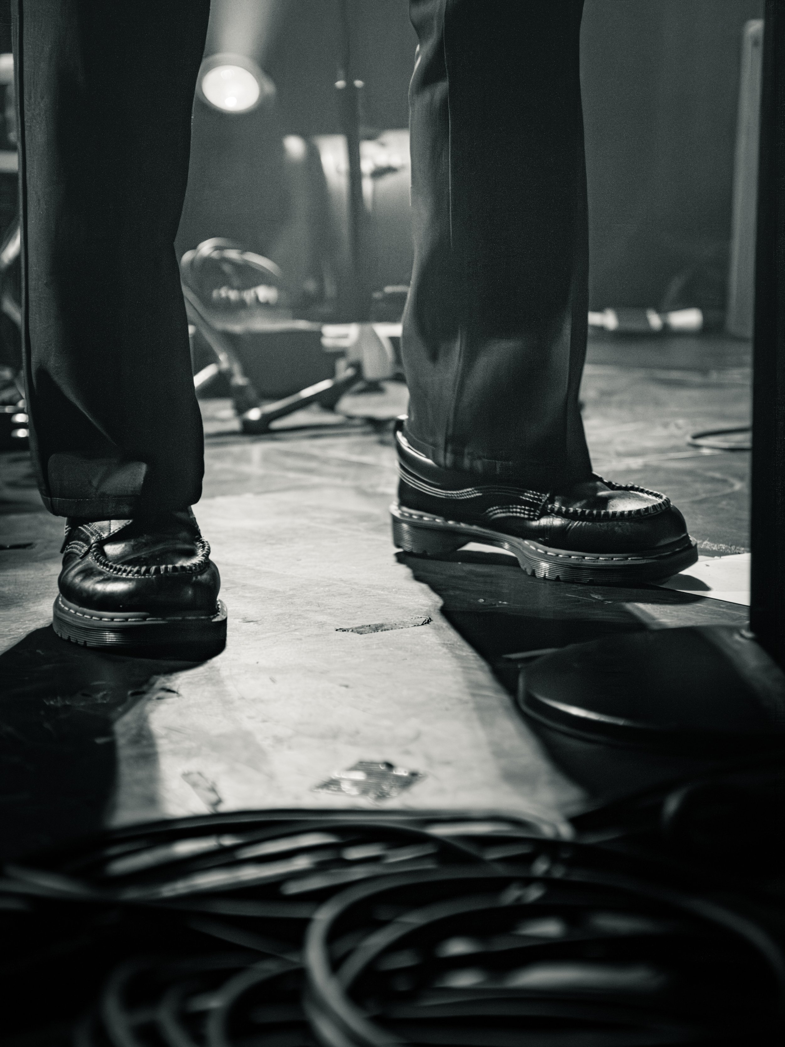 Close-up of a person wearing black leather shoes and black pants, standing on a wooden floor with wires scattered in the foreground, and blurred objects in the background.