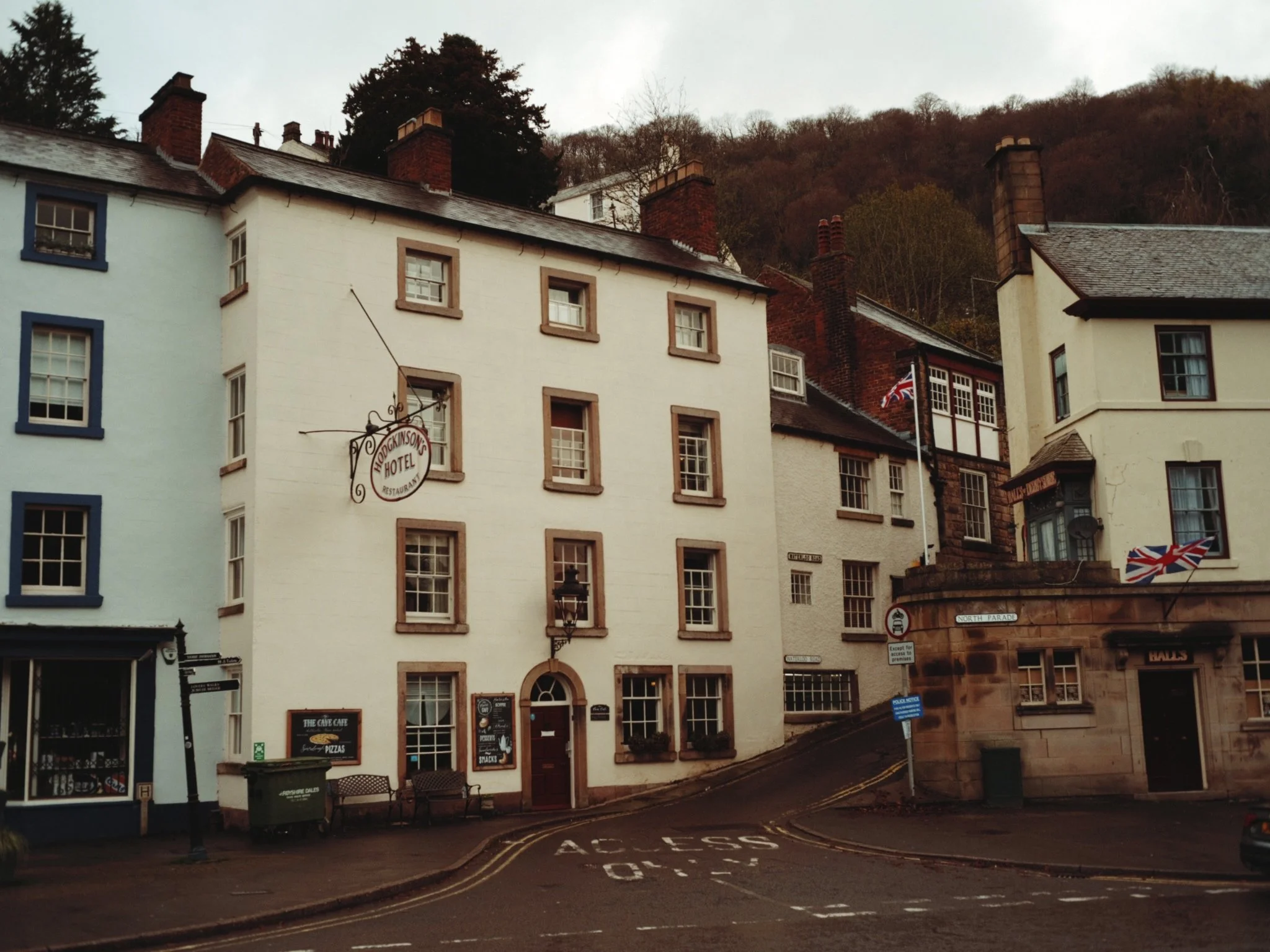 Street scene featuring a white hotel building with sign reading 'Hodgkinson's Hotel', adjacent buildings, Union Jack flags, and a hill with trees in the background.