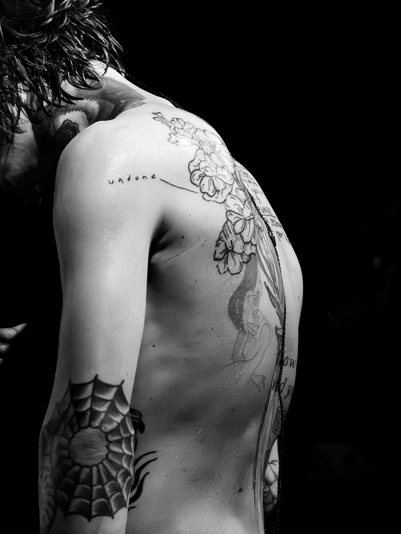A black and white photo of a person with wet hair, showing multiple tattoos on their shoulder and upper arm, including a spider web on the elbow, flowers on the shoulder, and outlines of other tattoos.