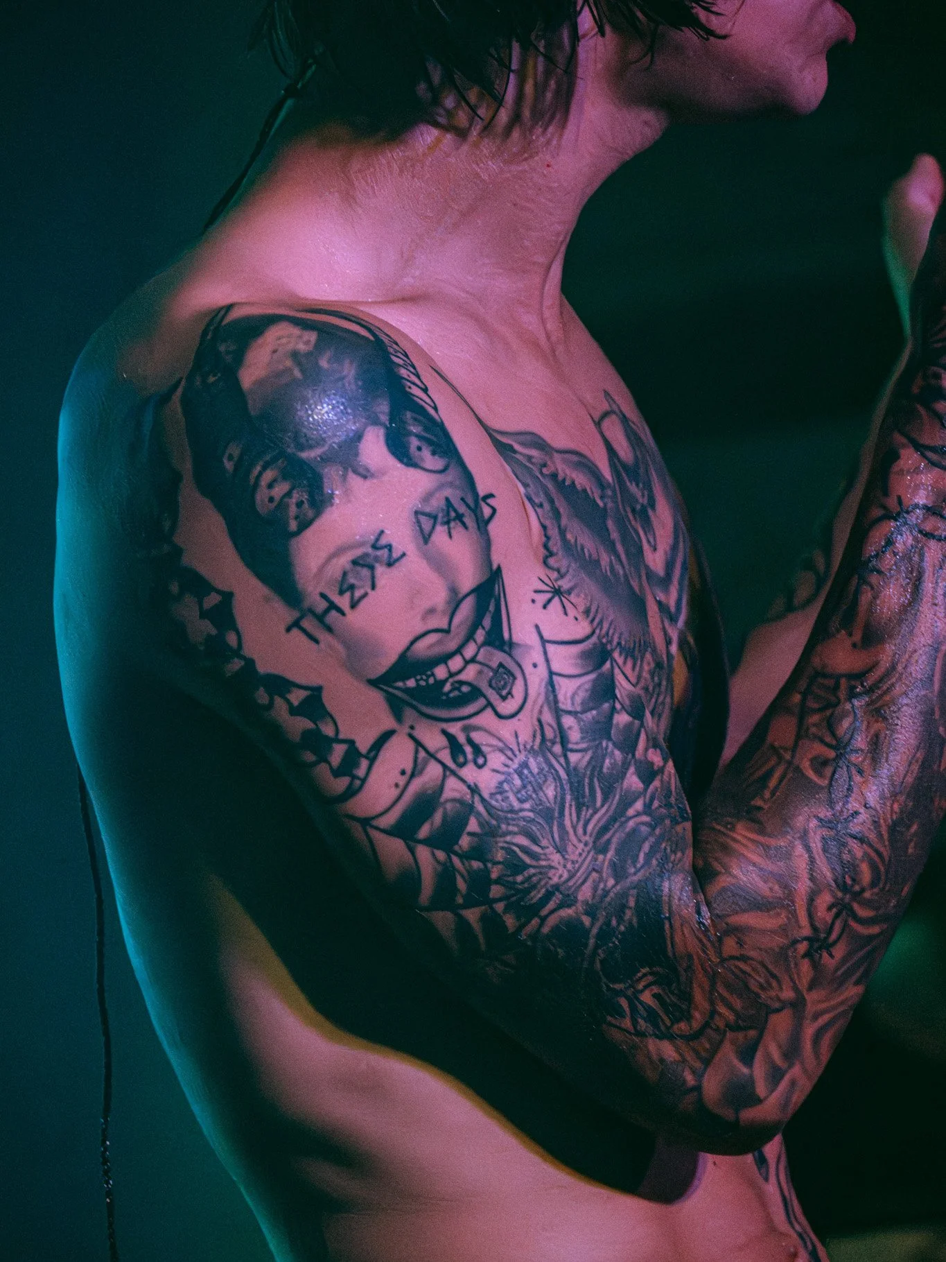 Close-up of a person's arm and shoulder with tattooed skin, showing the profile of a face and the phrase 'These Days' on the chest. The person has curly dark hair and is in a low-light setting with colorful lighting.