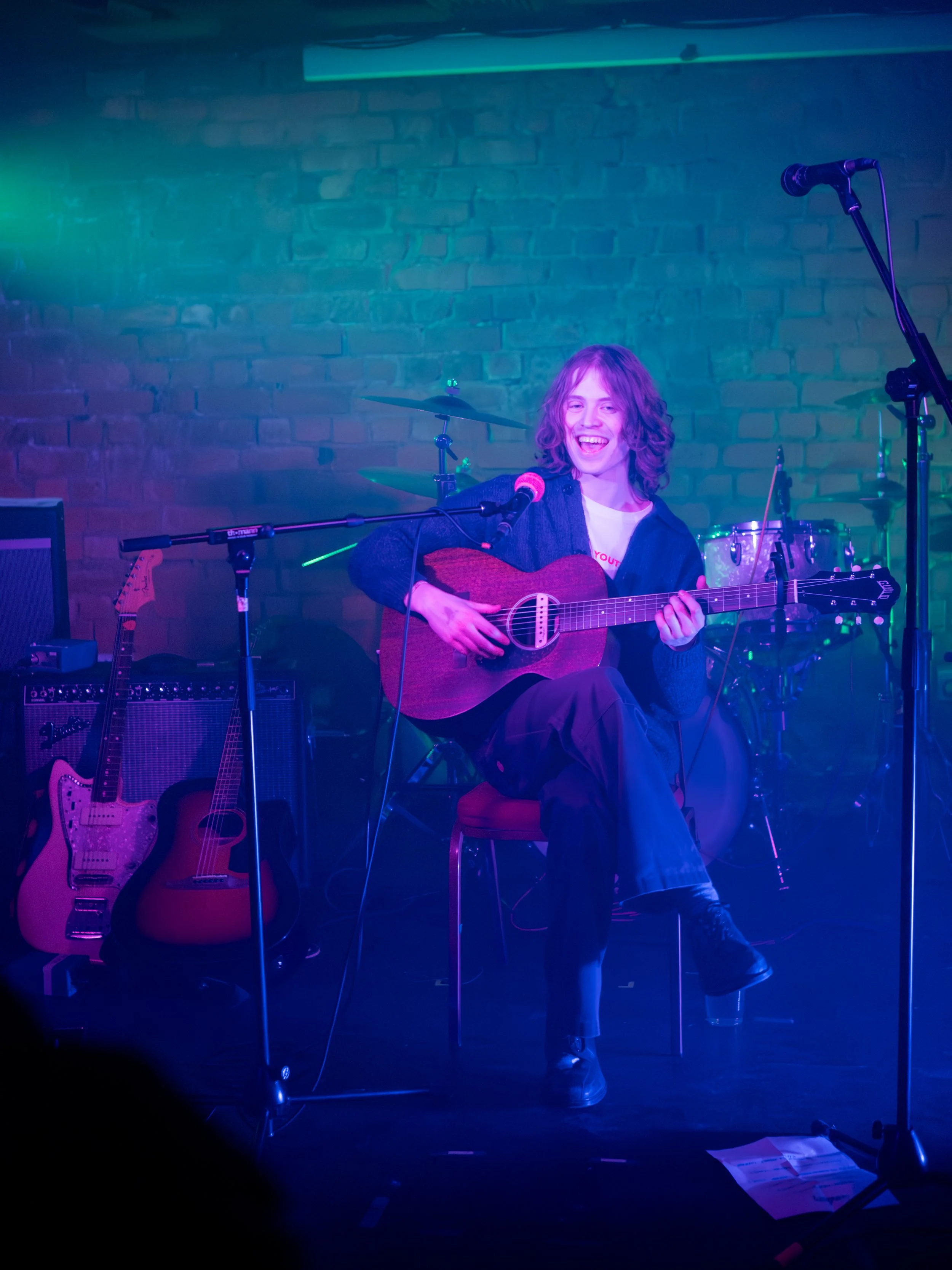 A woman playing guitar and singing into a microphone on stage, surrounded by musical instruments, with colorful stage lighting.