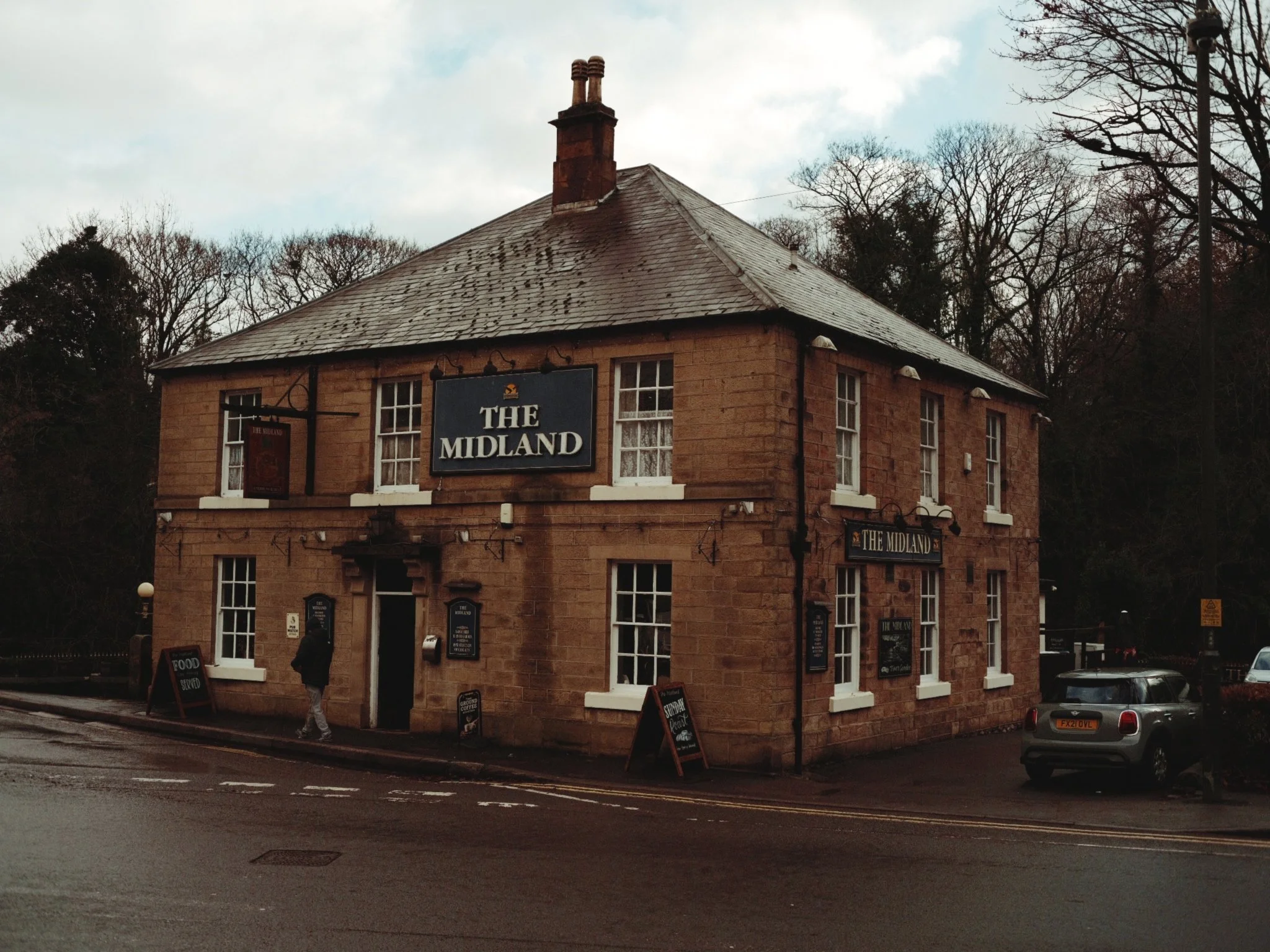 A two-story brick building with a sign reading 'The Midland,' with several windows, located on a street with parked cars and a pedestrian walking by.