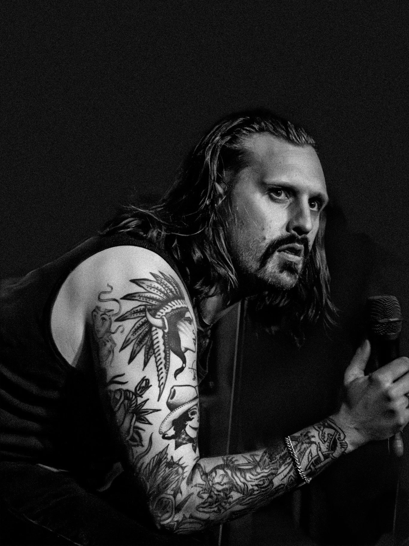 A man with long hair and tattoos on his arms holding a microphone in a dark setting.