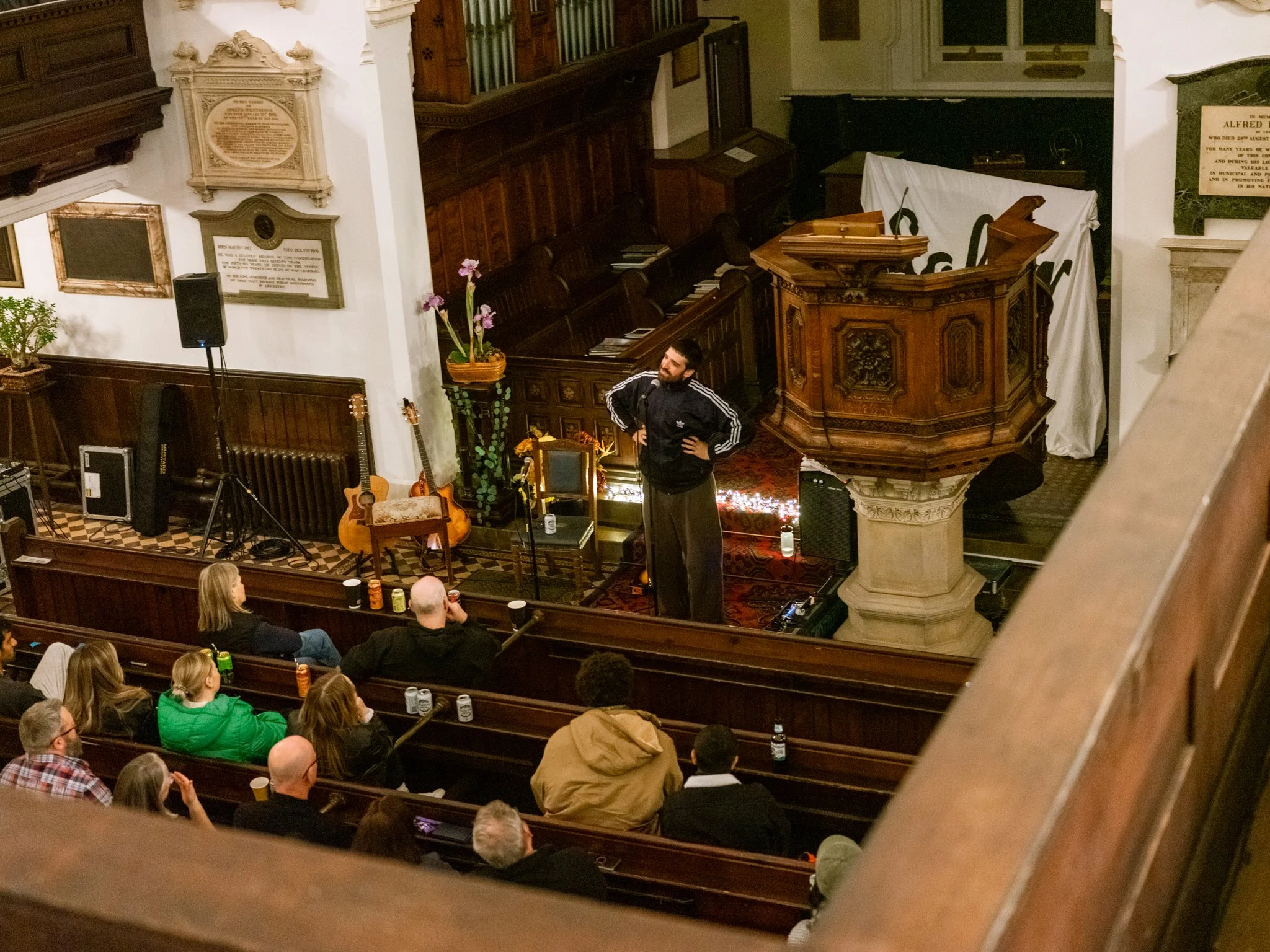 A man standing on a stage in a church, speaking or performing to an audience seated in pews. The stage has musical instruments, including guitars, and a microphone stand. The church is decorated with wooden panels, plaques, and plants.