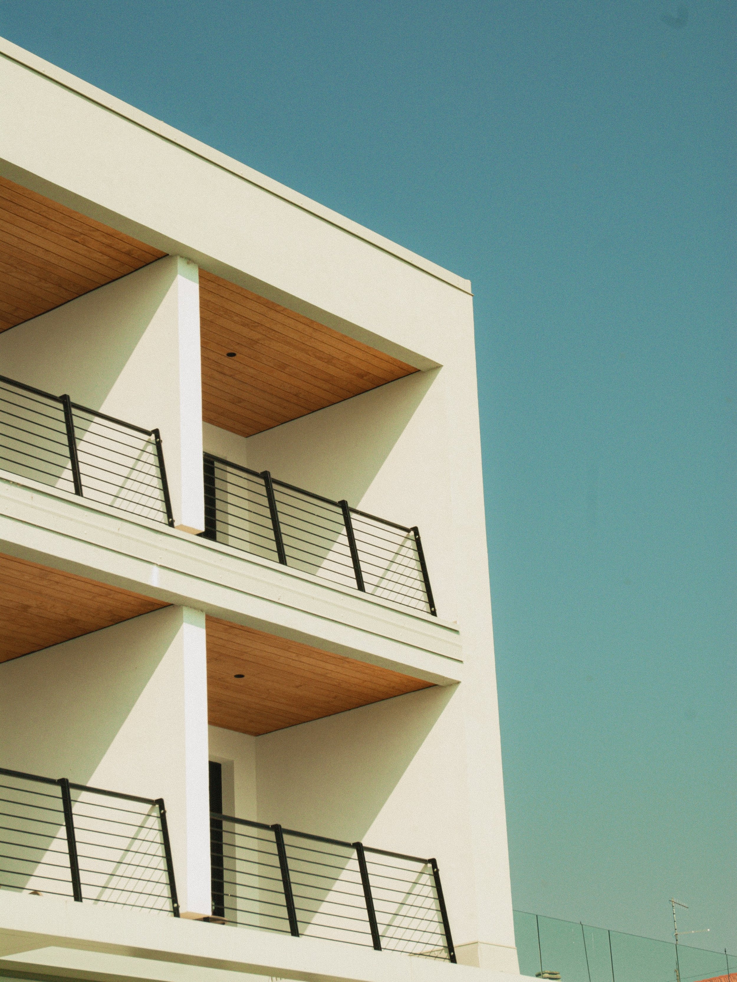 Close-up of a modern, white apartment building with balconies featuring black railings and wooden ceilings, against a clear blue sky.
