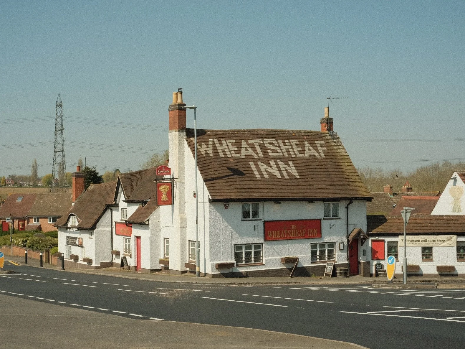 A white building with a brown tiled roof that has the words 'WHEATSHEAF INN' painted on it. There are signs and windows on the building, with a street in front and clear sky above.