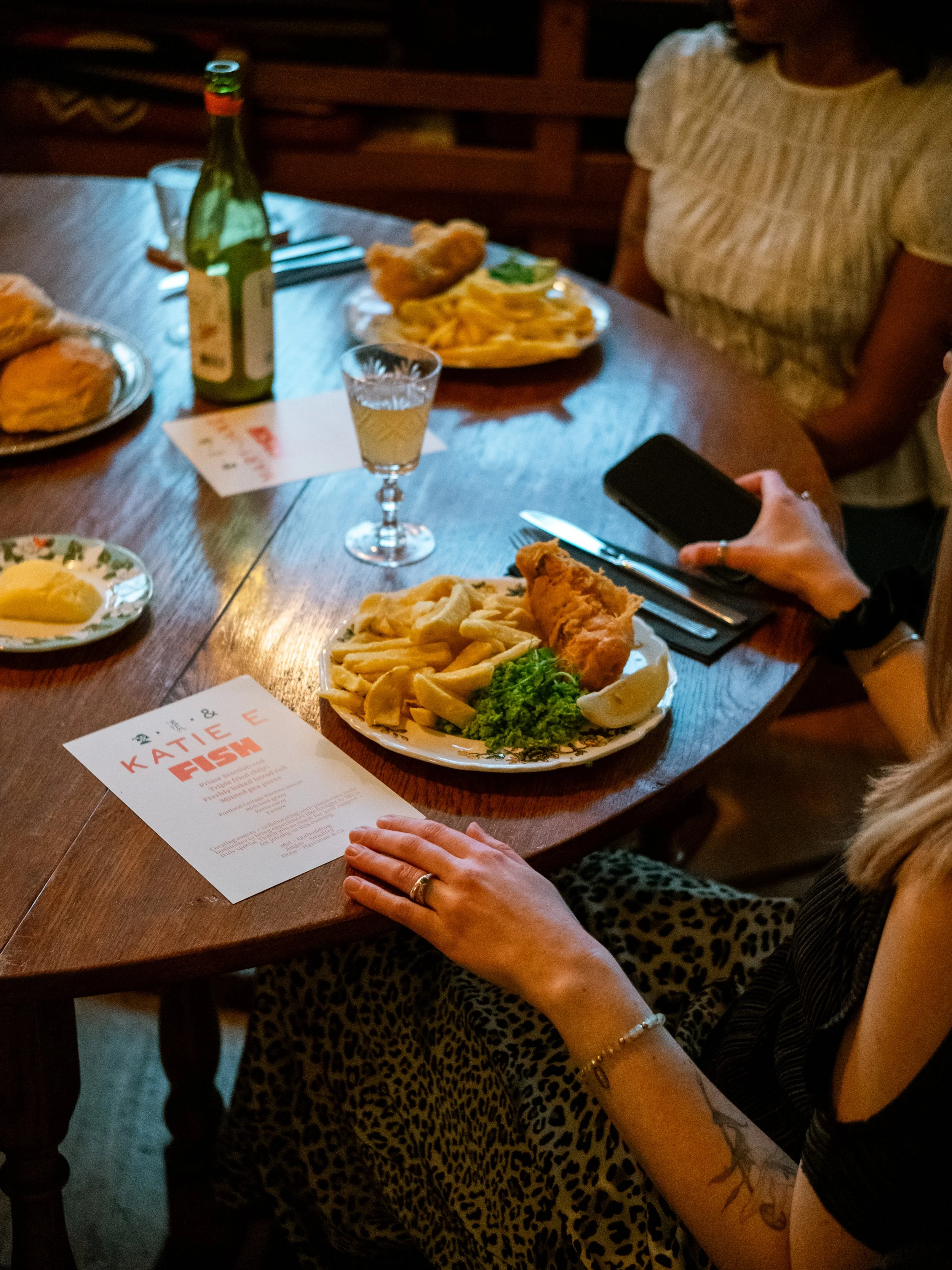 People dining at a wood table with plates of fish and chips, a glass of white wine, and a beer bottle in a cozy restaurant setting.