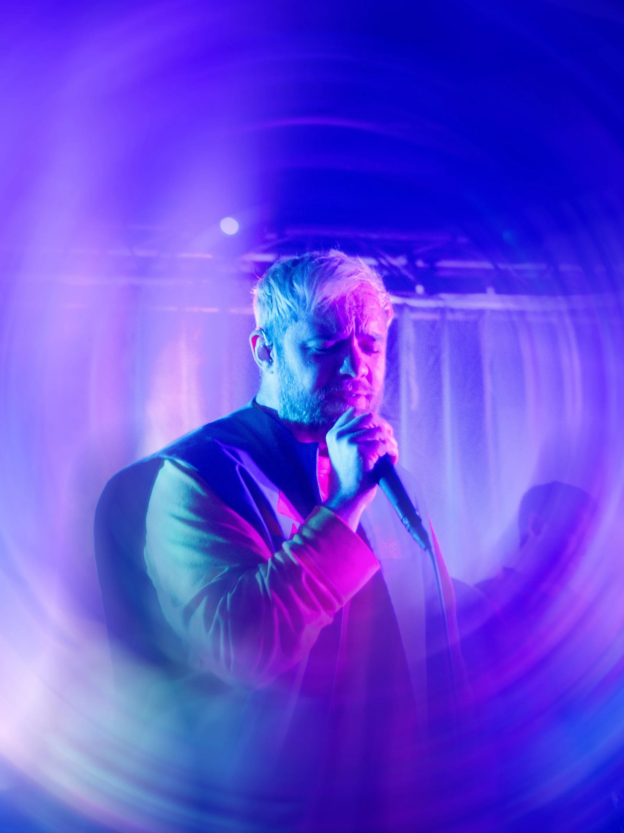 A male singer with blonde hair and a beard performing on stage in colorful purple and blue lighting, holding a microphone and closed eyes.