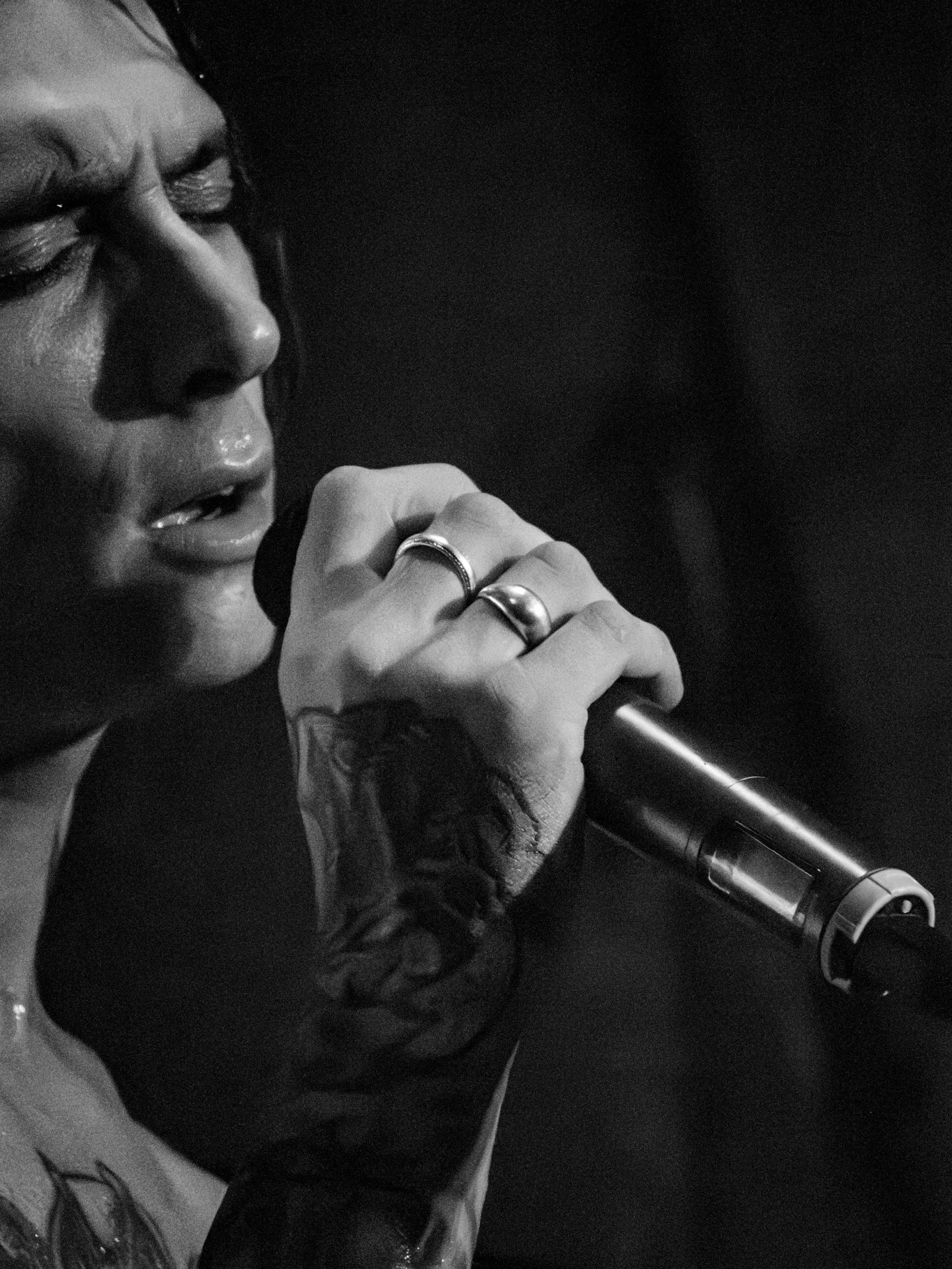 Close-up of a woman singing into a microphone, with tattoos and rings on her hand, in a black-and-white photo.