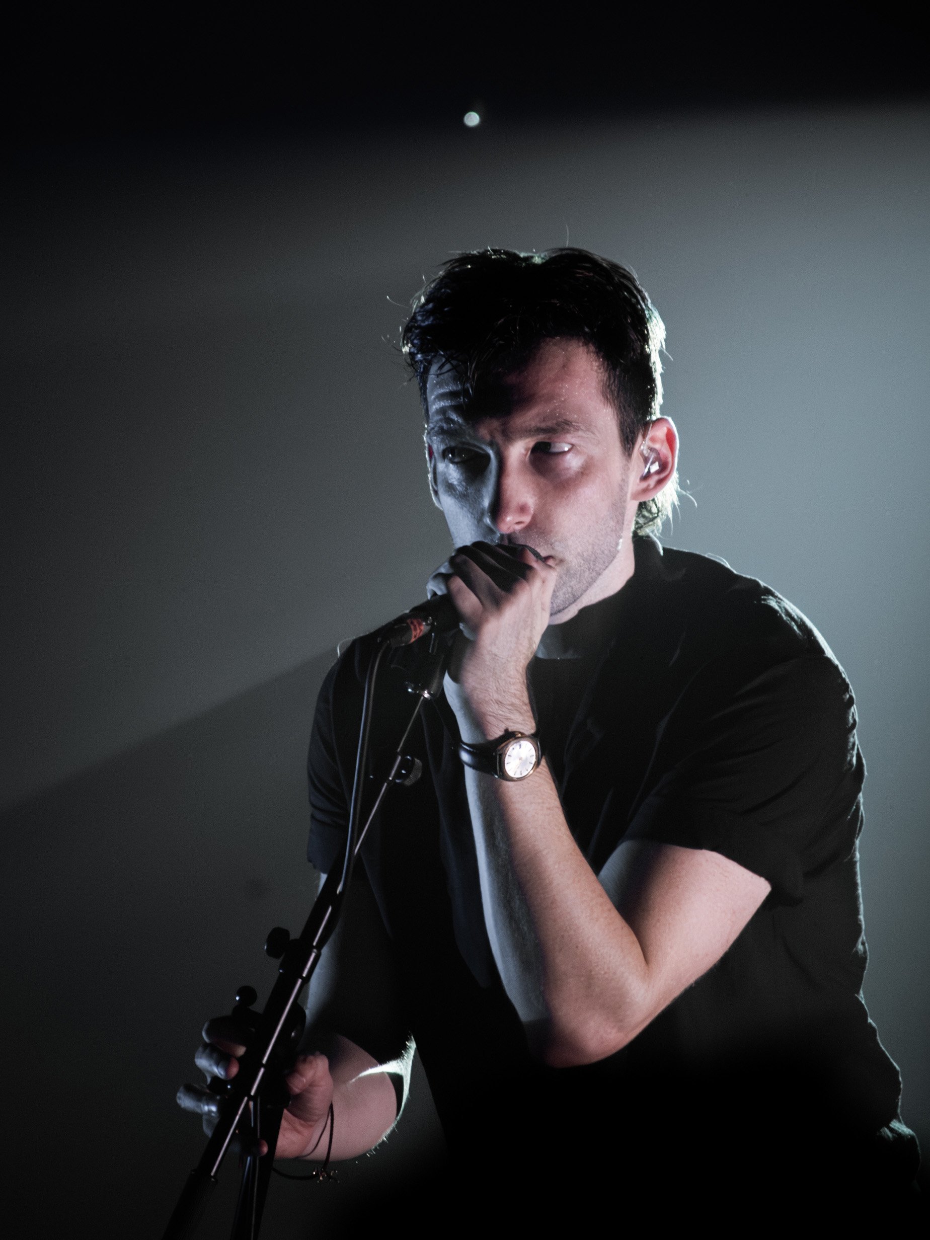 A male singer performs on stage, holding a microphone close to his mouth, with dramatic lighting creating shadows on his face and body.