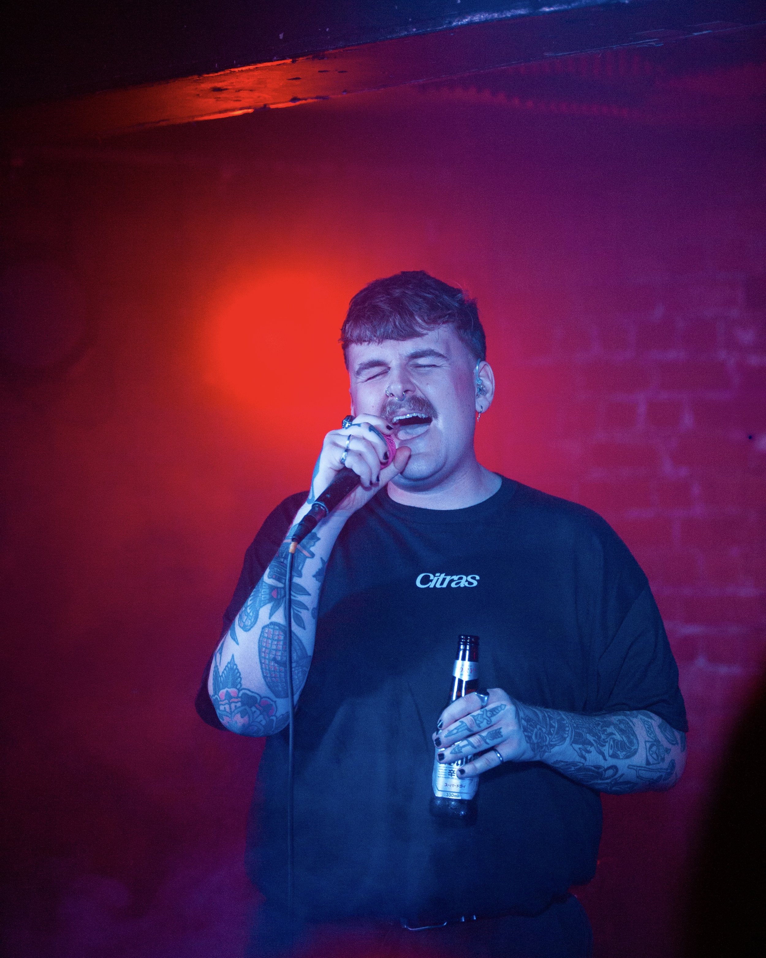 A young man singing into a microphone while holding a beer bottle, with colorful stage lighting and a brick wall background.