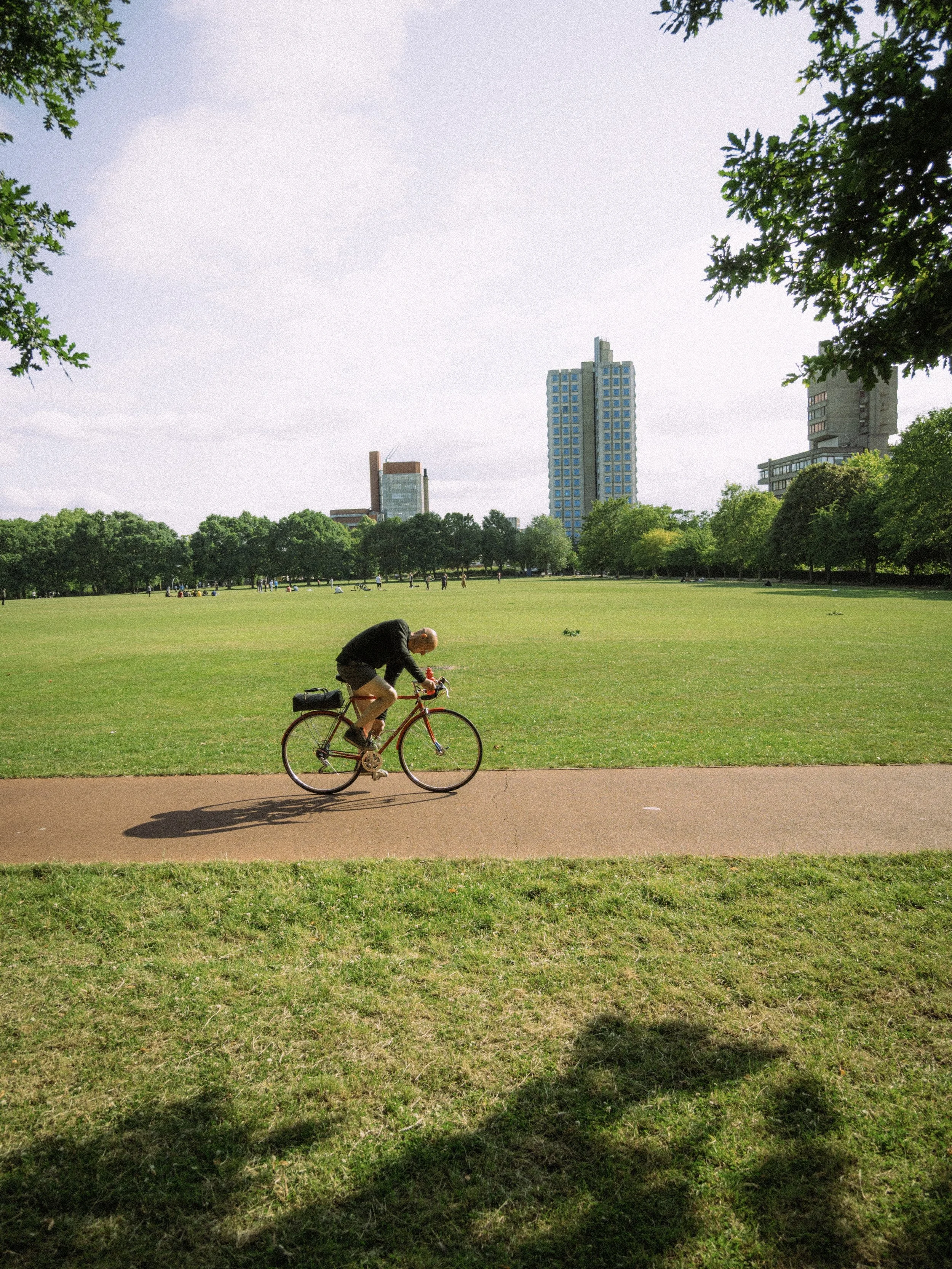 A person riding a red bicycle on a paved path in a green park with a city skyline in the background.