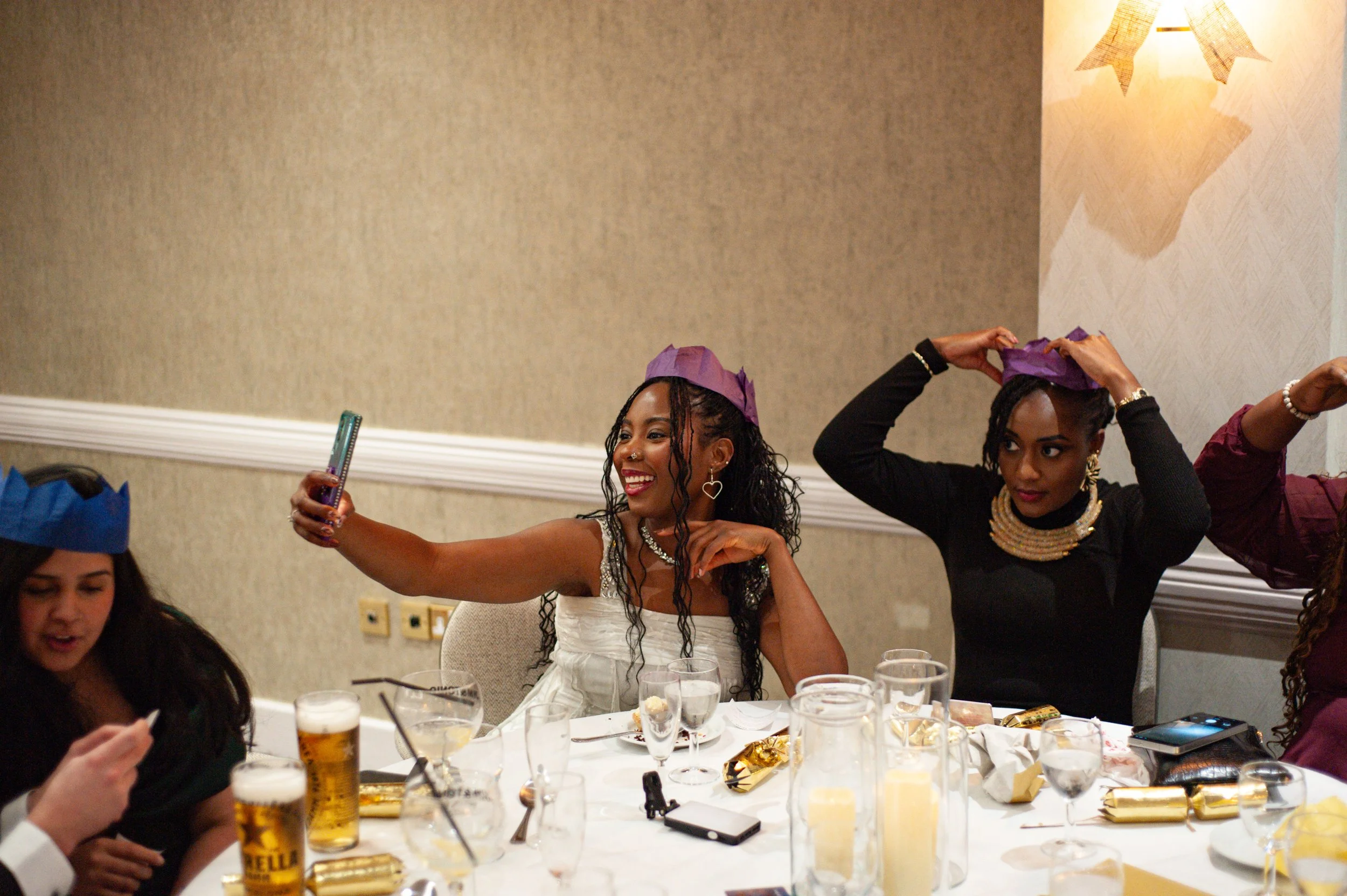 Women wearing purple and blue paper hats, taking a selfie and adjusting their hats at a celebration event with glasses and drinks on the table.