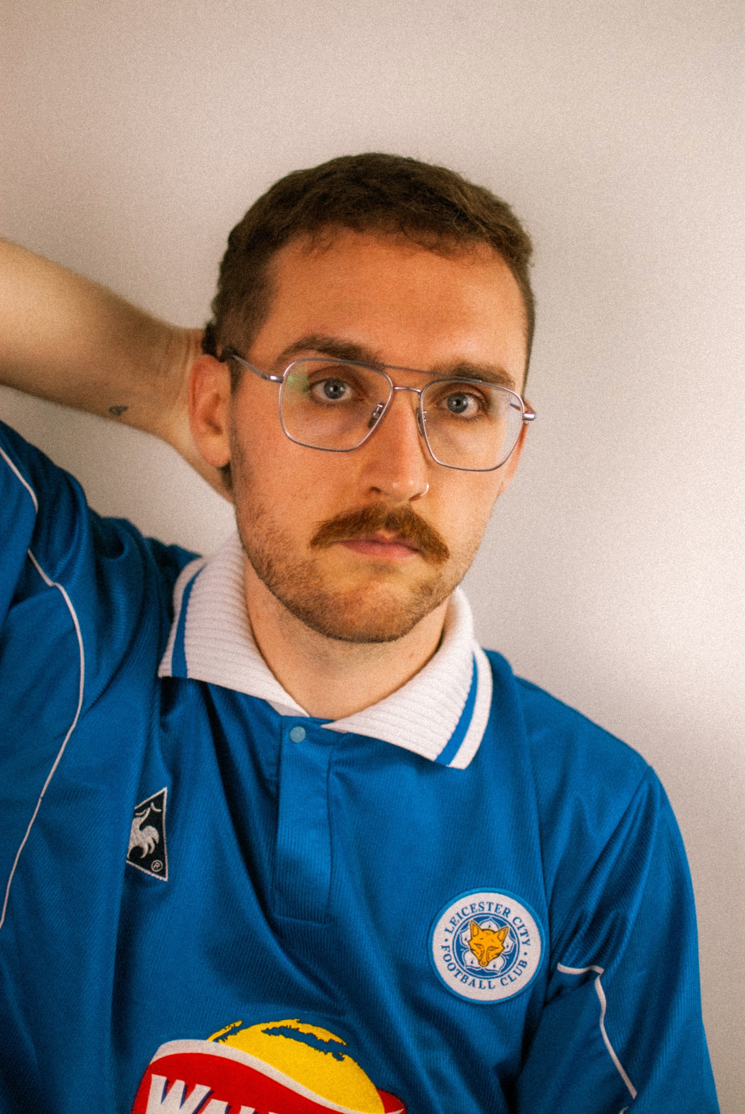 A man with glasses and a mustache, wearing a blue Leicester City football jersey, posing with one hand behind his head against a plain light-colored wall.