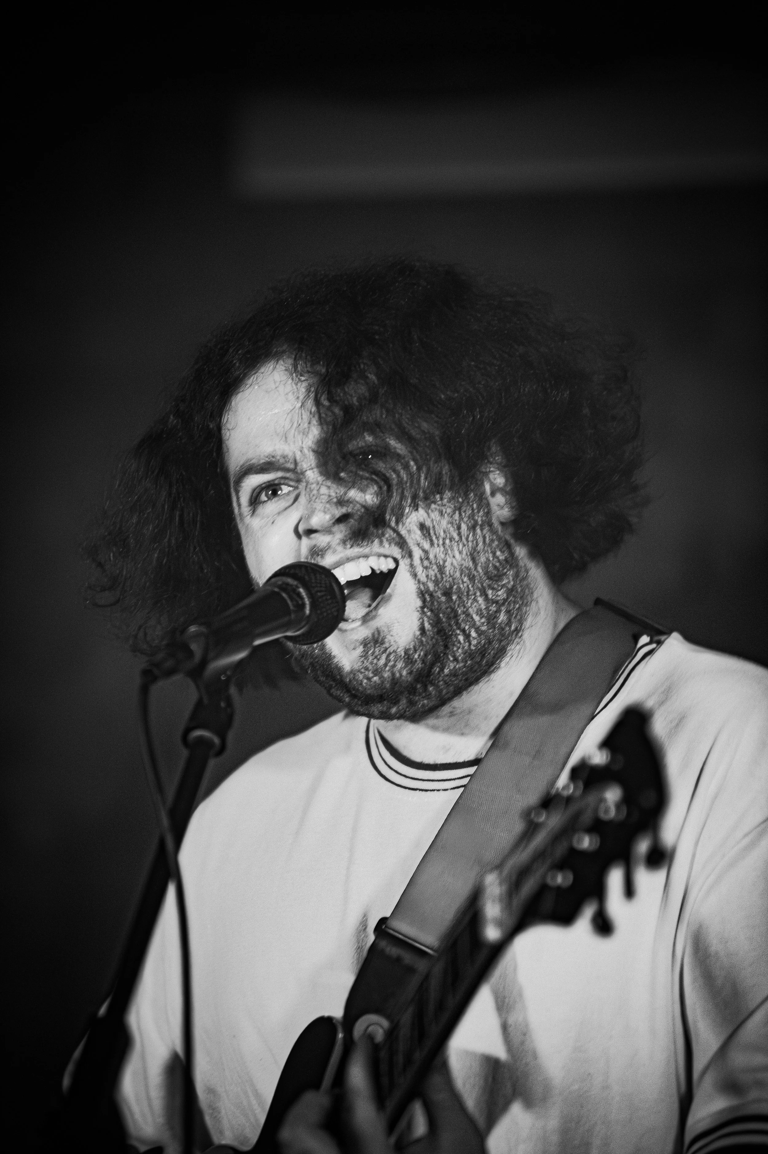 A black-and-white photo of a man singing into a microphone while playing an electric guitar. He has curly, disheveled hair and a beard, wearing a light-colored shirt with dark stripes around the collar.