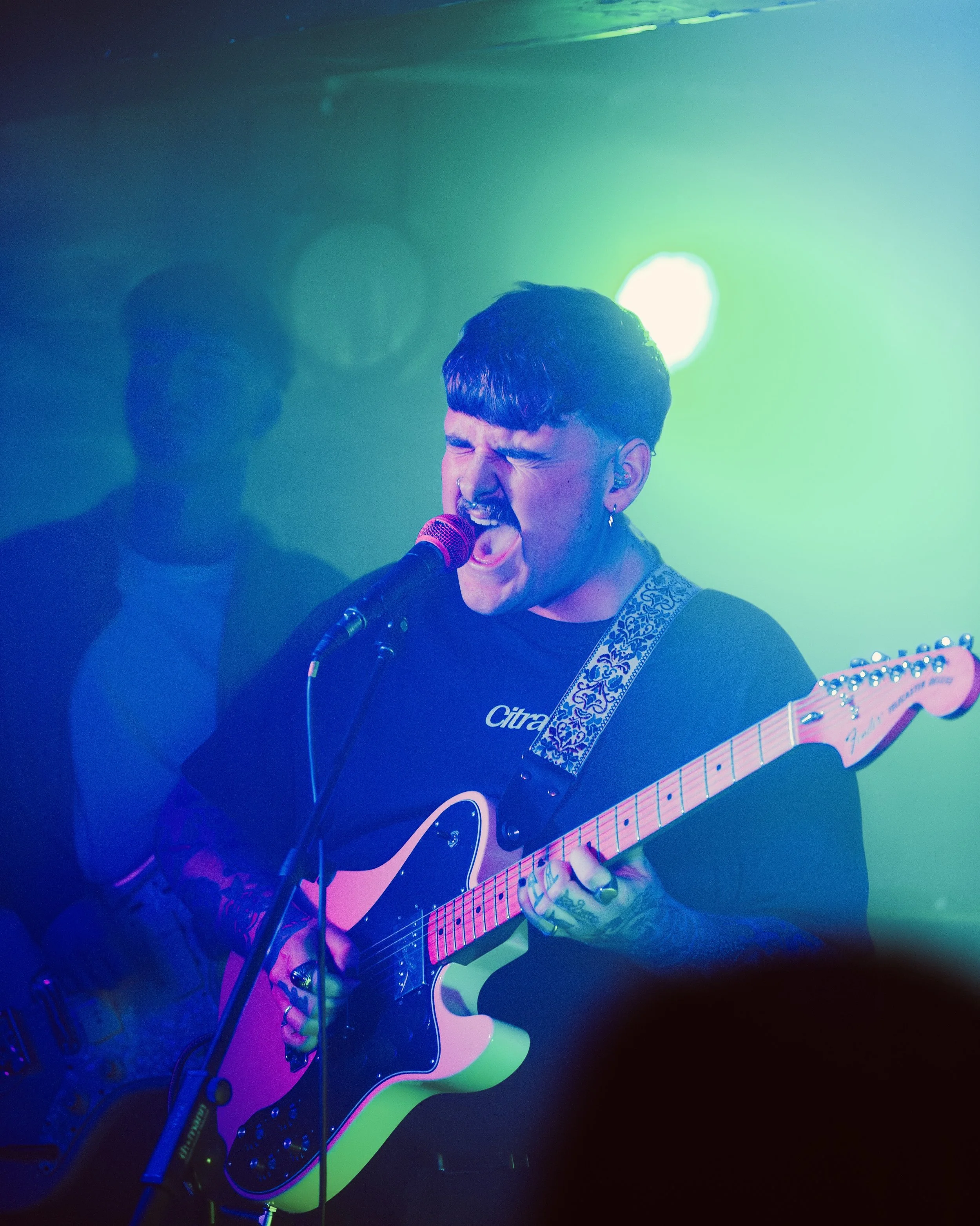 A musician passionately singing into a microphone while playing an electric guitar on stage with colorful lighting.