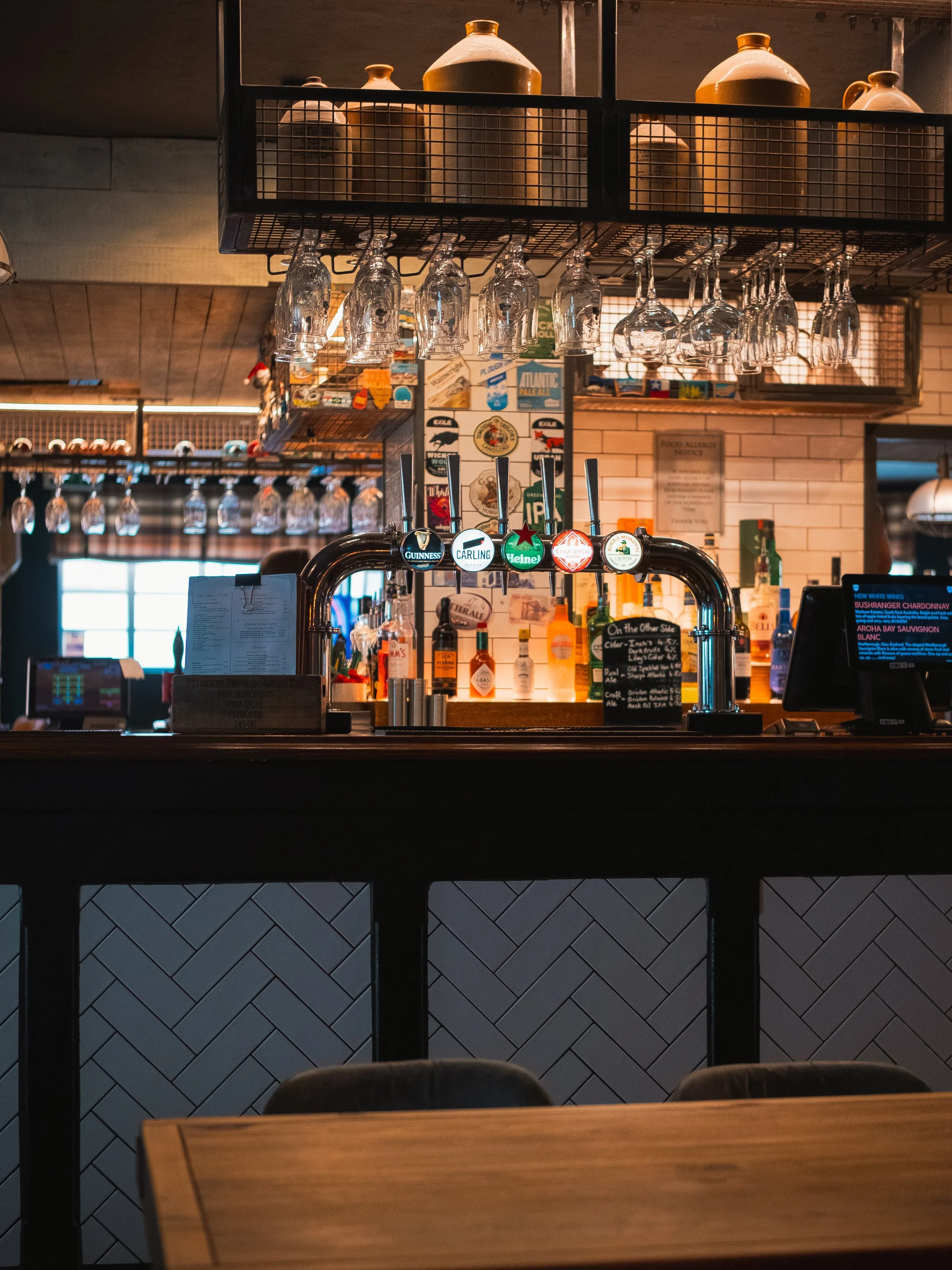 Interior of a bar with hanging wine glasses and beer taps, bottles of alcohol, and a chalkboard menu.