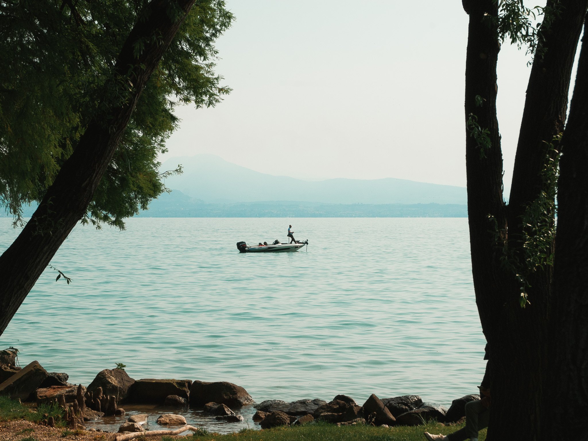 A person on a boat on a large body of water, with mountains in the background and trees framing the scene.