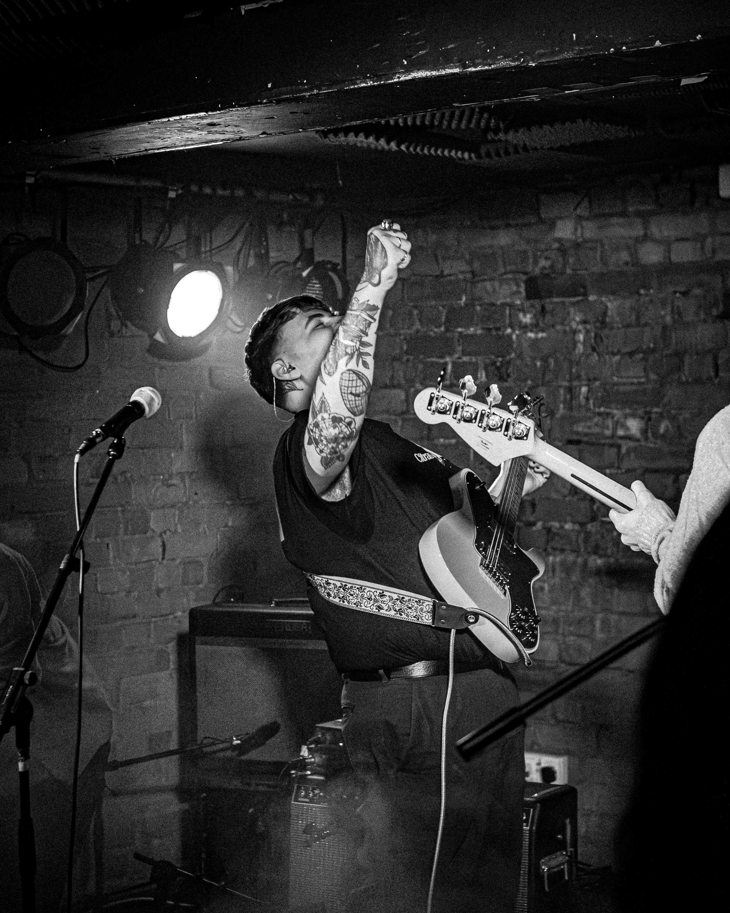 A woman with tattoos singing passionately and playing an electric guitar on stage with a brick wall background, in a black and white photo.