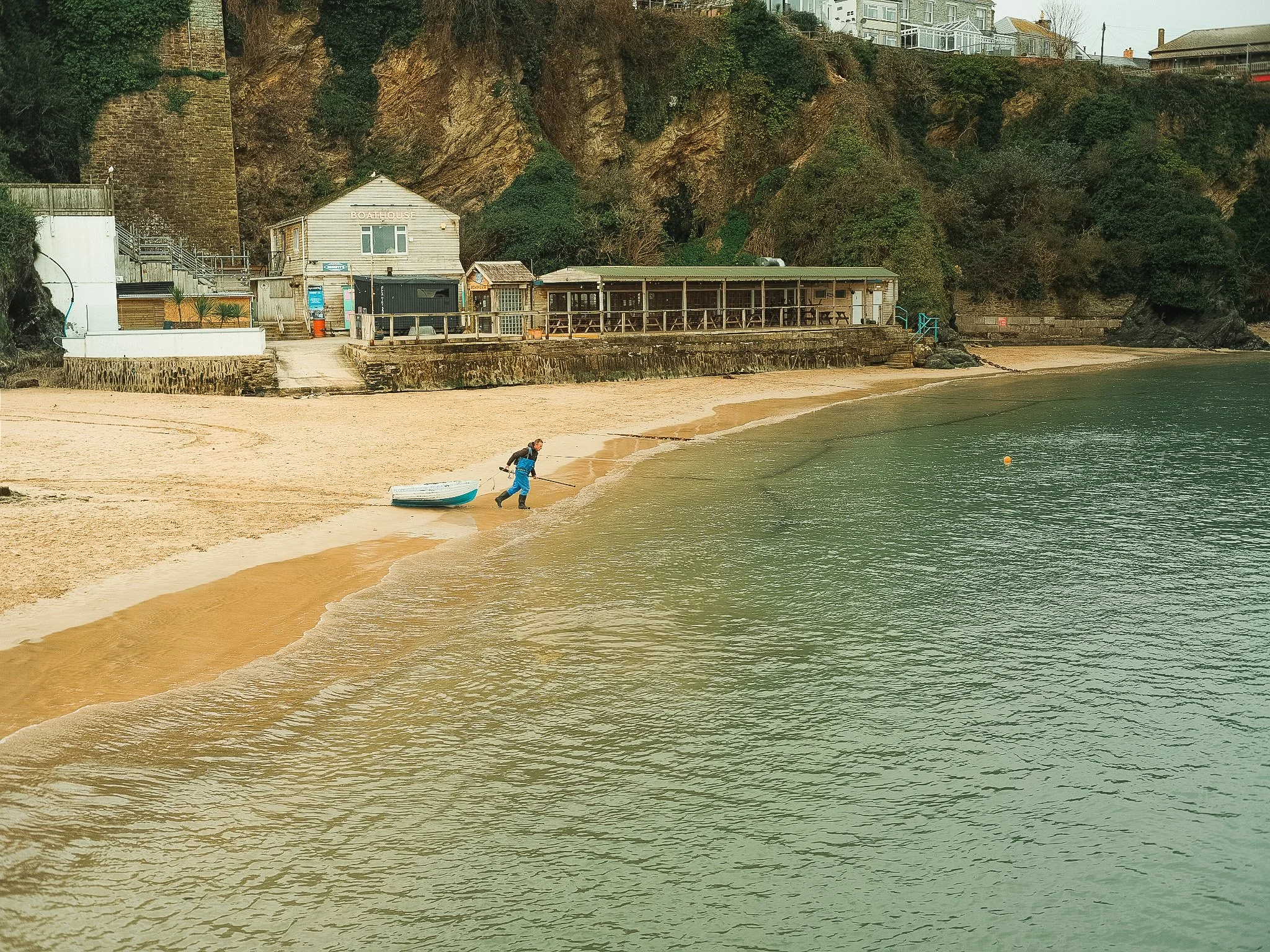 Person pulling a small boat along the sandy beach near the shoreline with houses and cliffs in the background.
