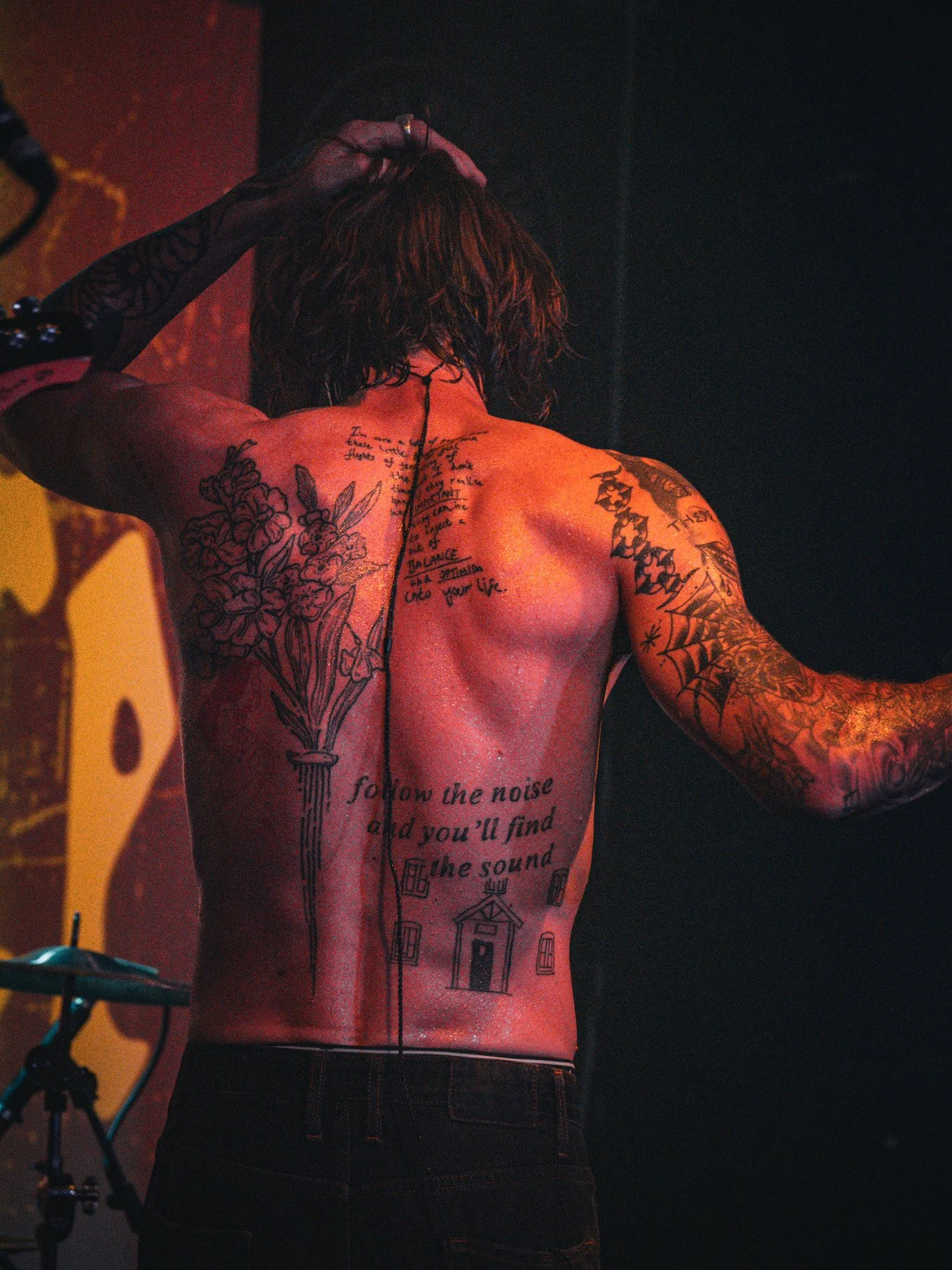 A shirtless man with tattoos on his back and arms, standing on stage with his left arm raised and his right hand touching his head, with a dark background.