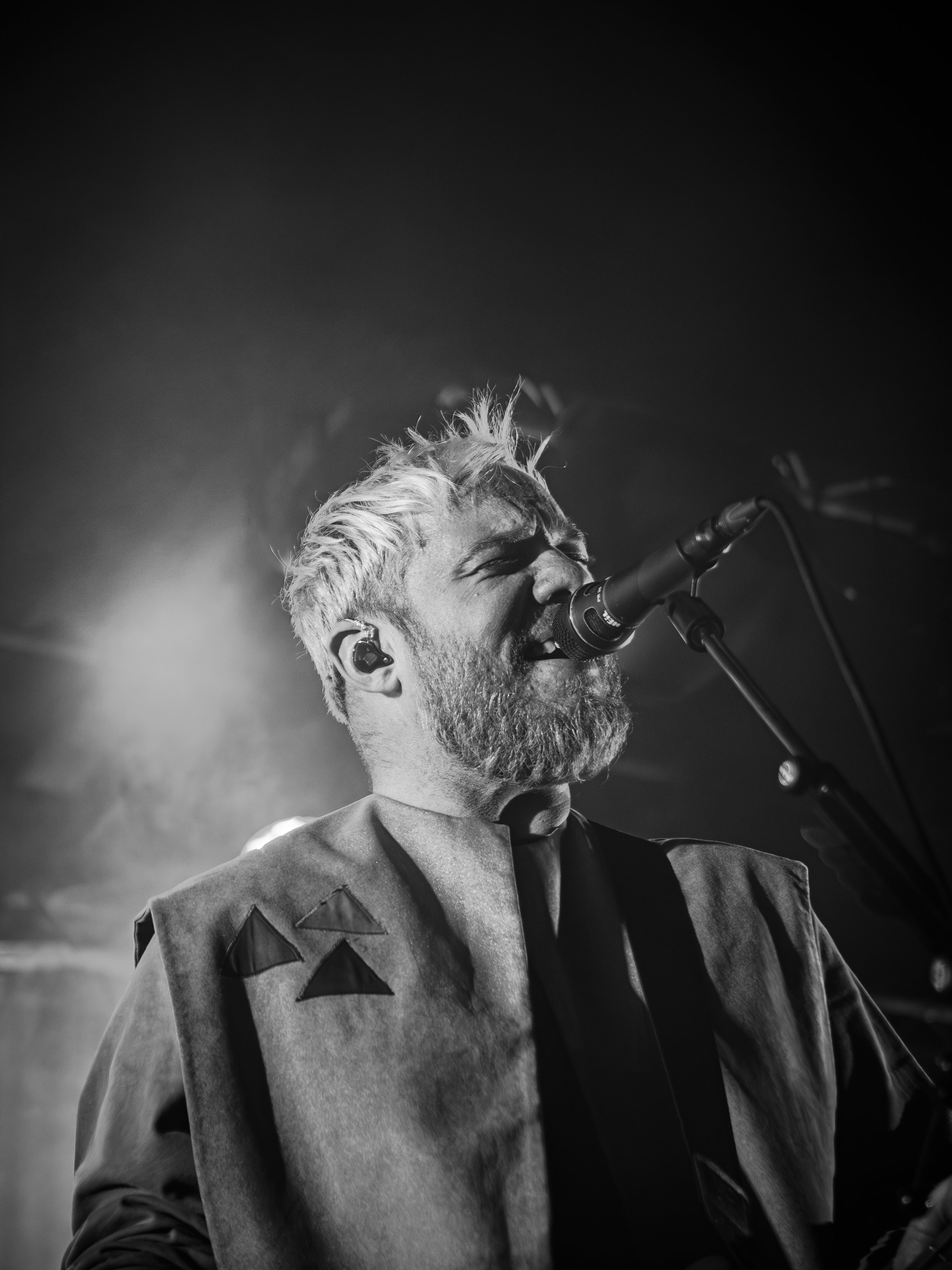 Black and white photo of a male singer with a beard singing into a microphone on stage, with closed eyes and a passionate expression.