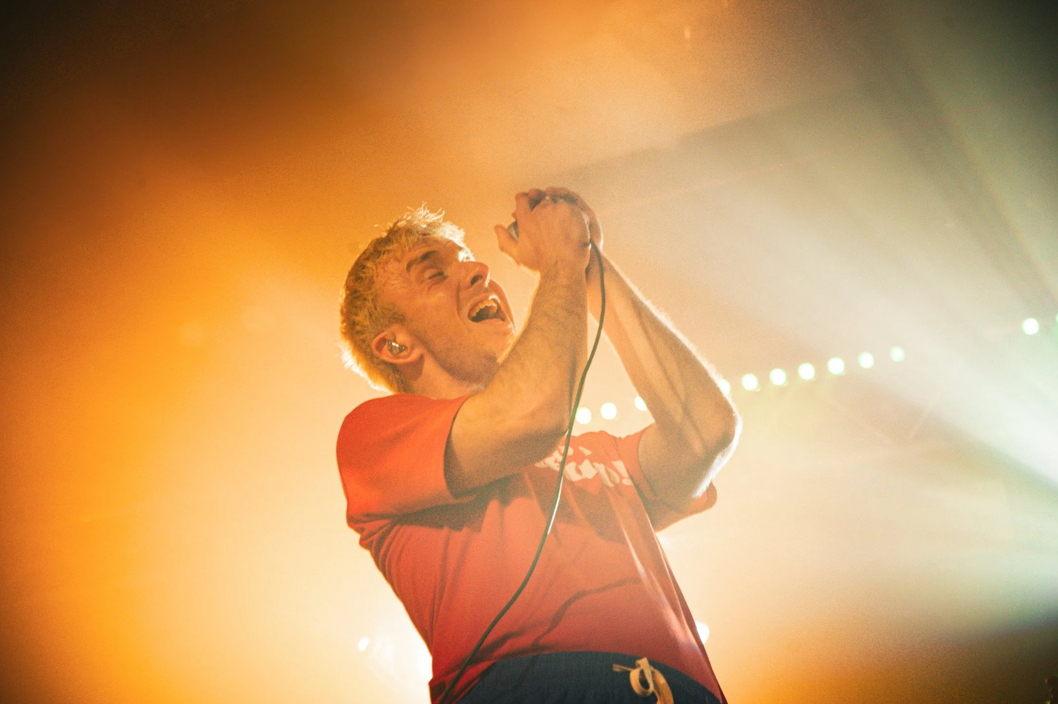 A male singer with blonde hair wearing a red shirt, passionately singing into a microphone on stage surrounded by colorful lighting.