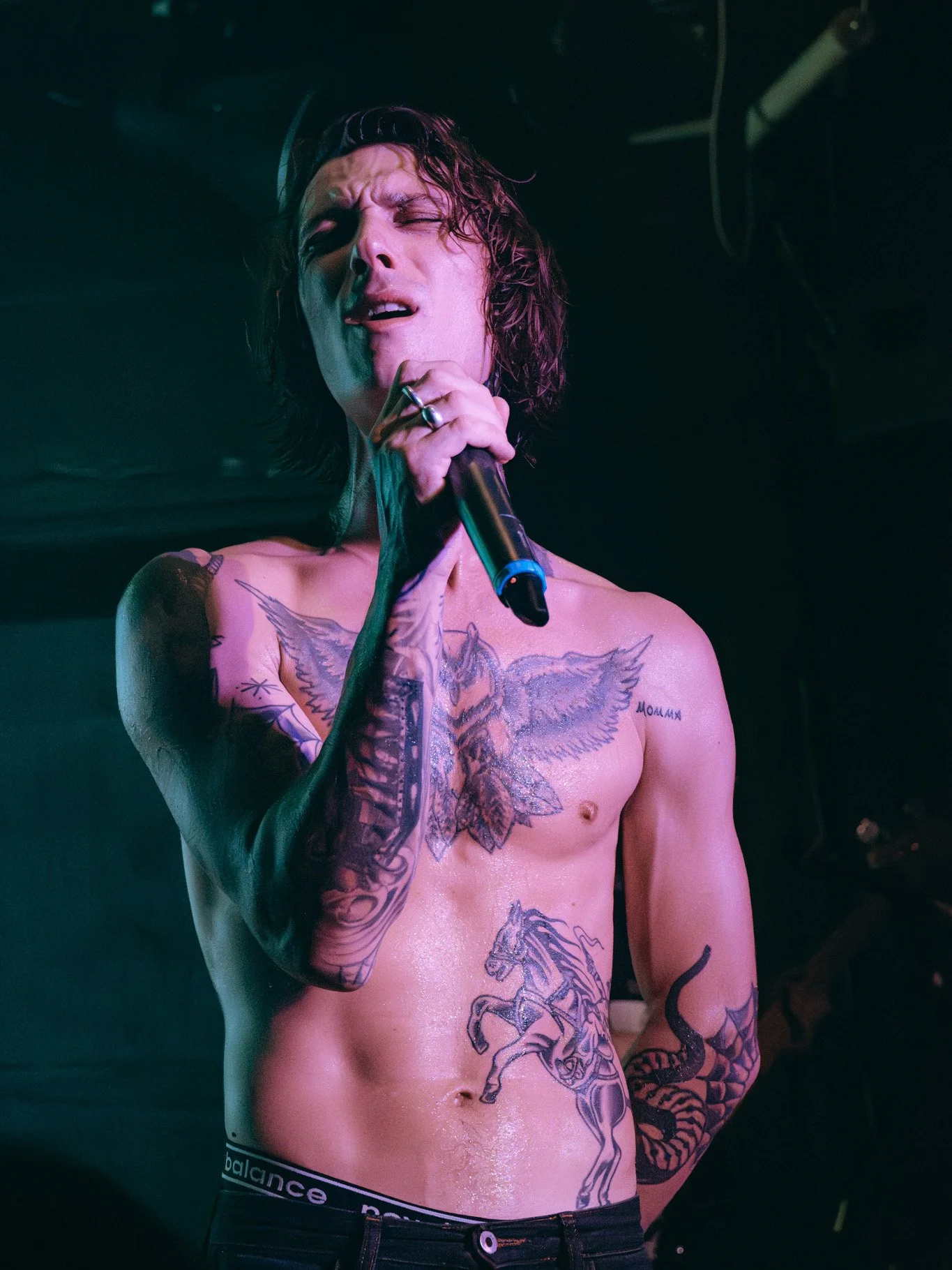 A shirtless man with long, dark, curly hair singing into a microphone, tattoos covering his chest and arms, with a dark background.