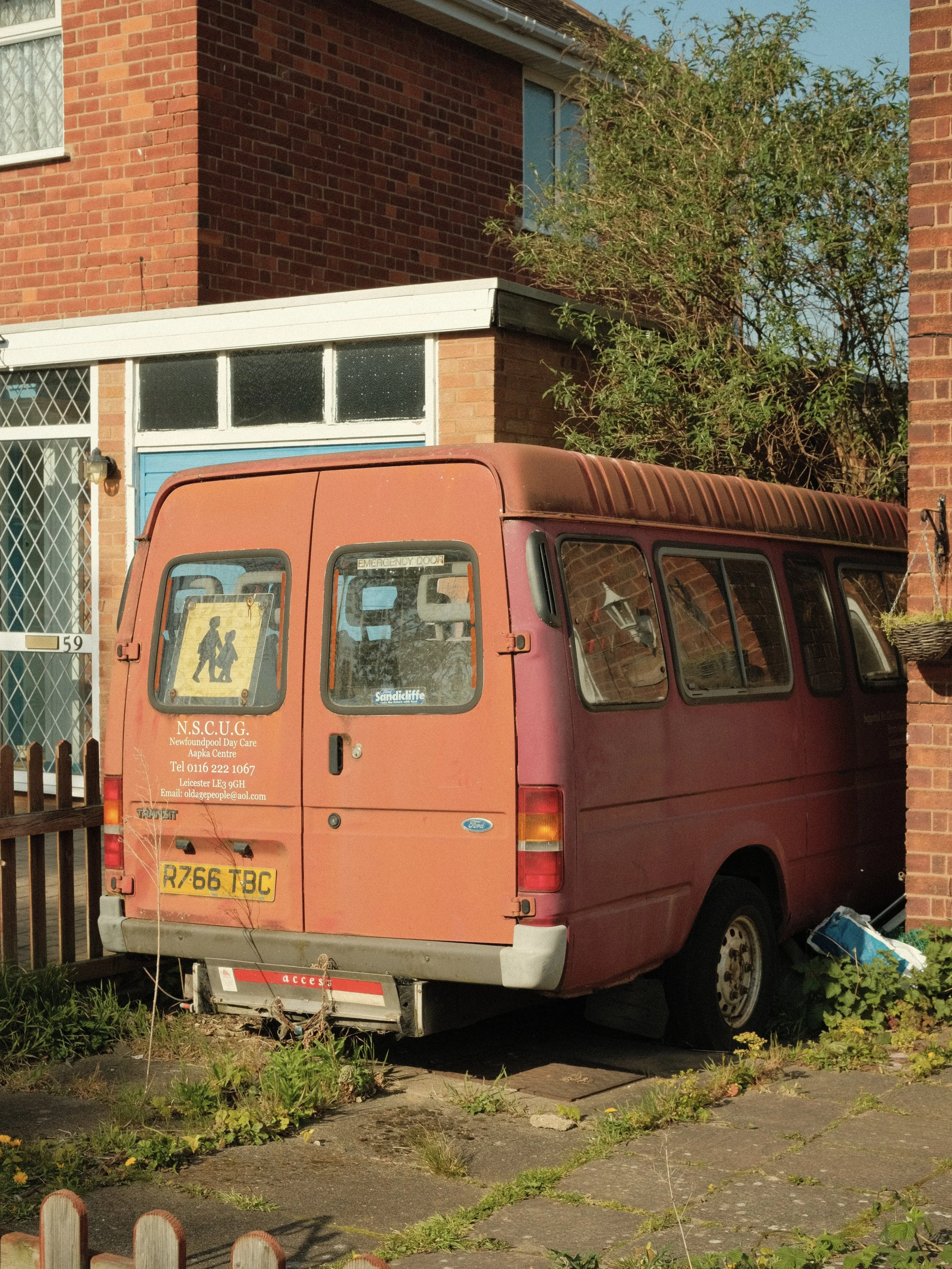 An old, red van parked on a concrete driveway next to a brick house with a fenced yard, some grass and weeds at the base of the van, and a large green tree behind the house.