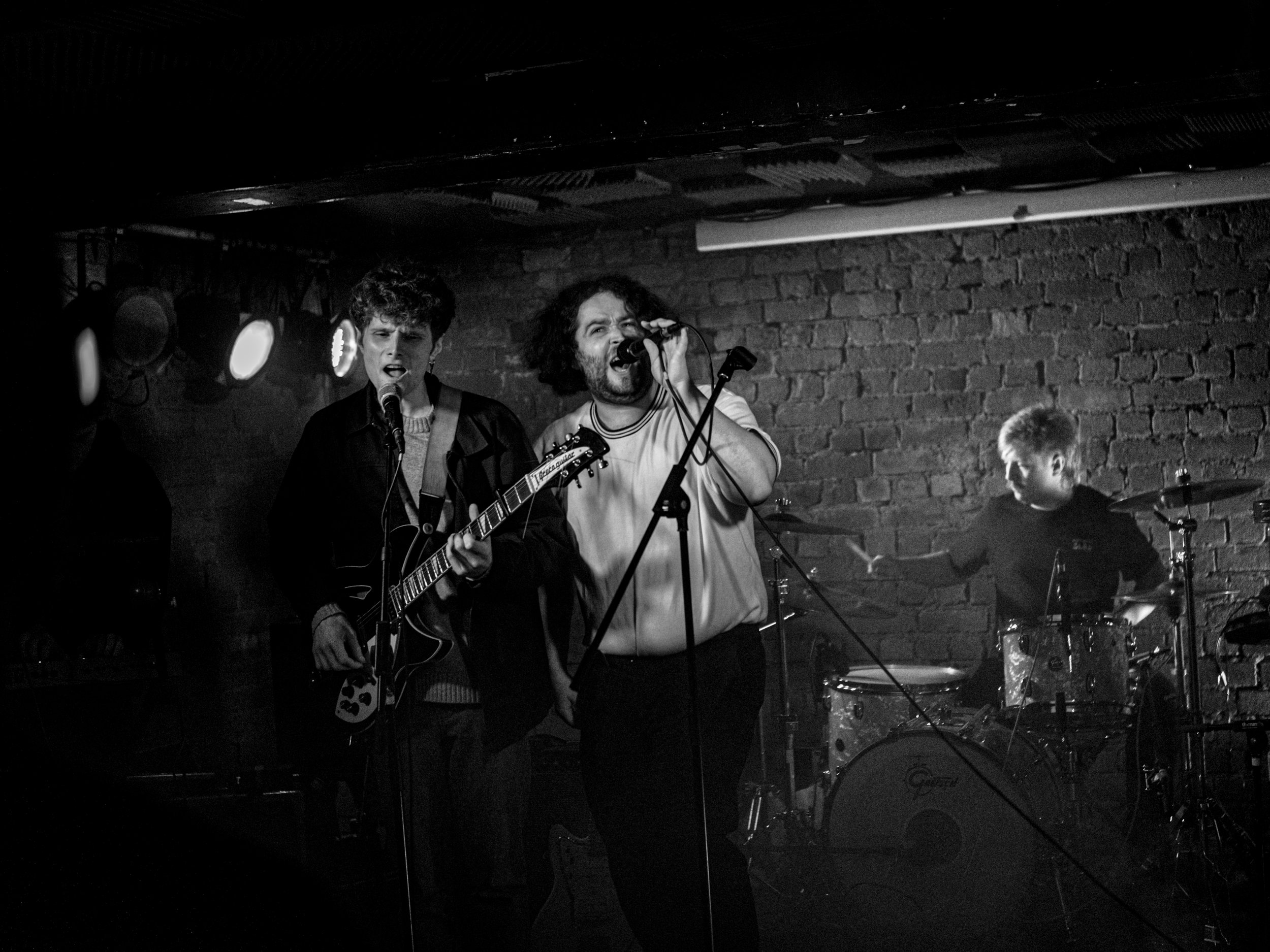 A black-and-white photo of a band performing on stage. The band includes a guitarist, a vocalist, and a drummer playing against a brick wall background. The guitarist is positioned on the left, singing into a microphone. The vocalist is in the center