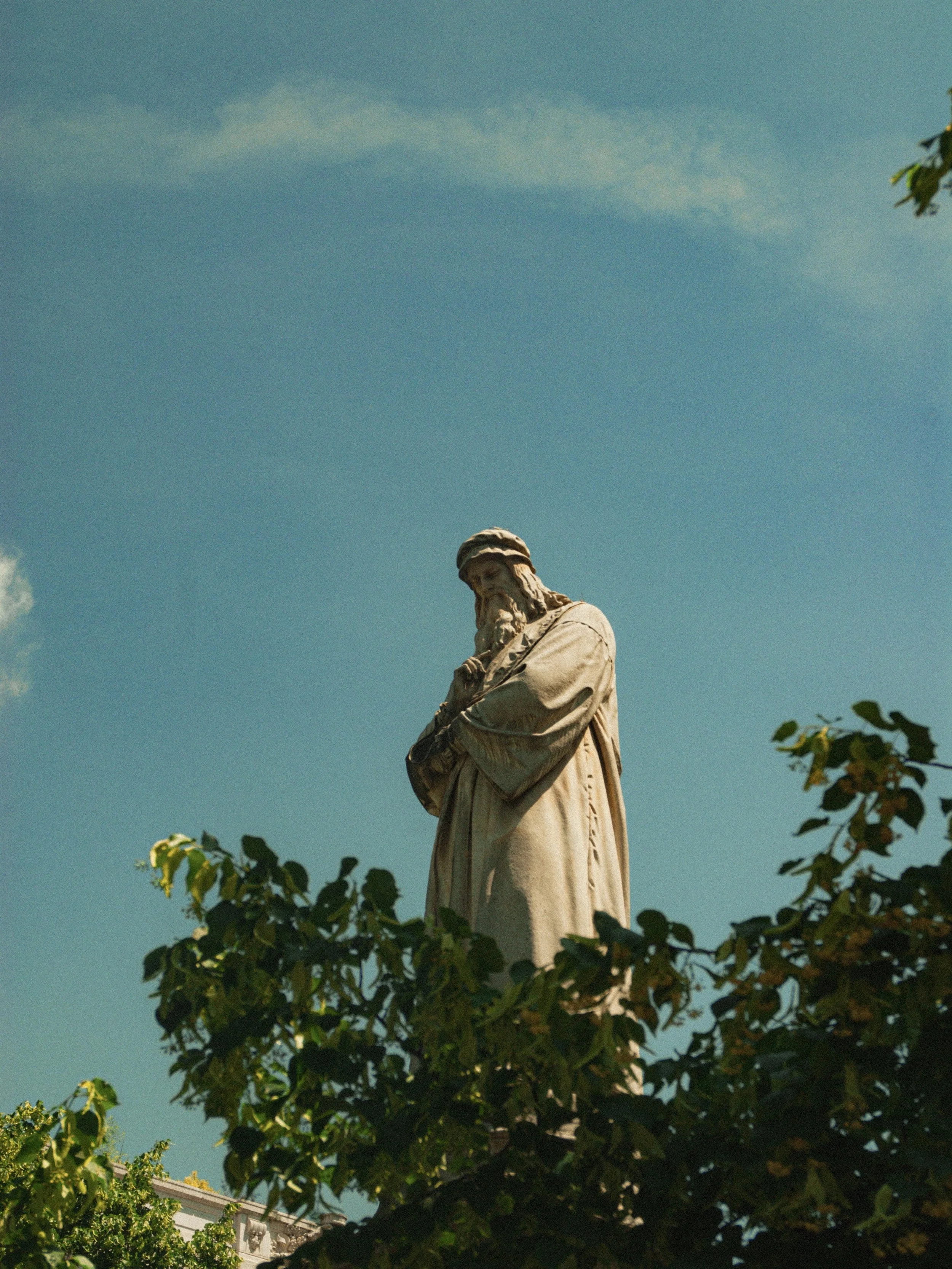 Statue of a man with a beard and head covering, surrounded by green trees, against a blue sky with a few clouds.