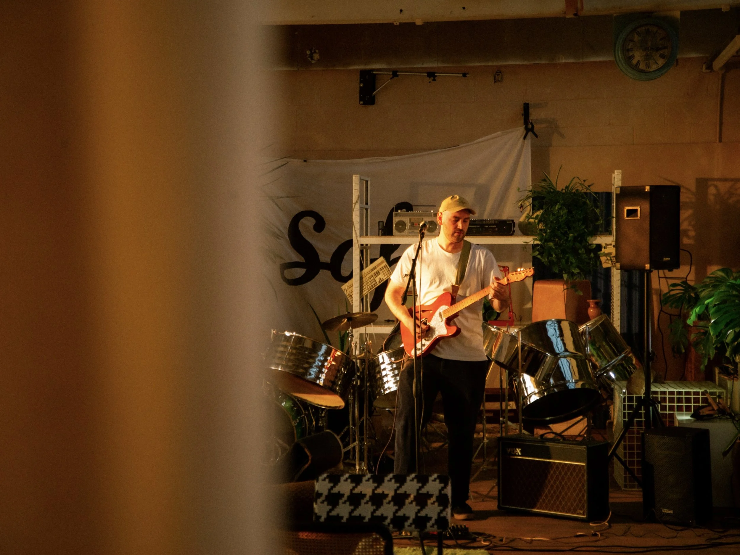 A musician playing an electric guitar on stage with musical equipment and plants, partially obscured by a blurred object in the foreground.